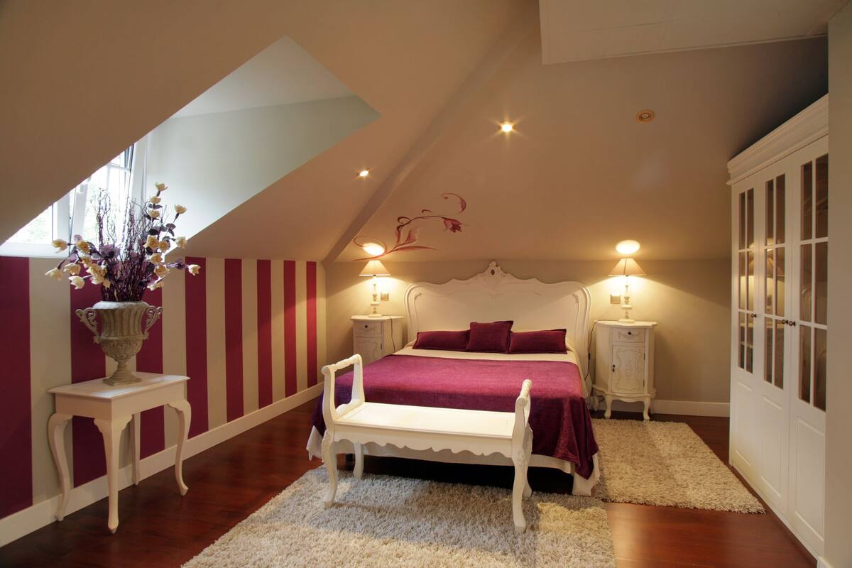 Un dormitorio con techo inclinado, una cama con cabecera blanca, dos mesitas de noche con lámparas, un banco blanco, un jarrón con flores y una ventana con vistas.
