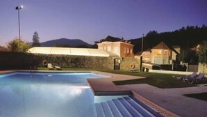 A swimming pool with a stone wall and a covered area, surrounded by houses and a mountain in the background.