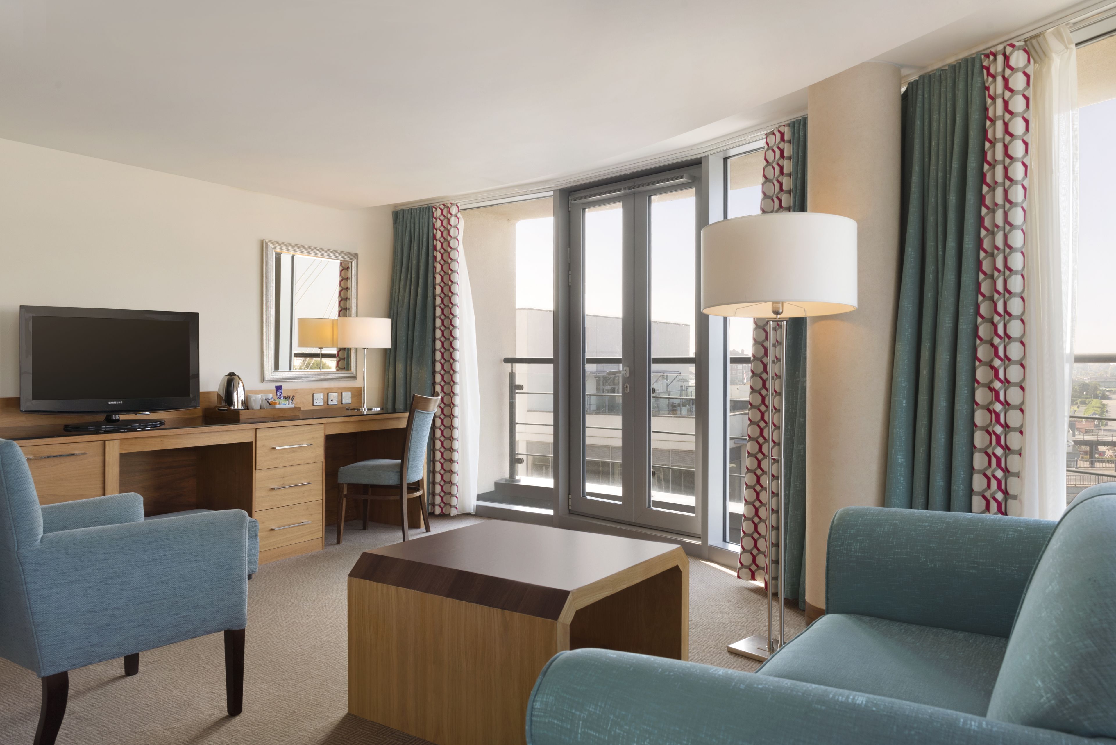 A hotel room with a flat-screen TV, a desk with a chair, a sofa, a coffee table, and large windows with curtains.