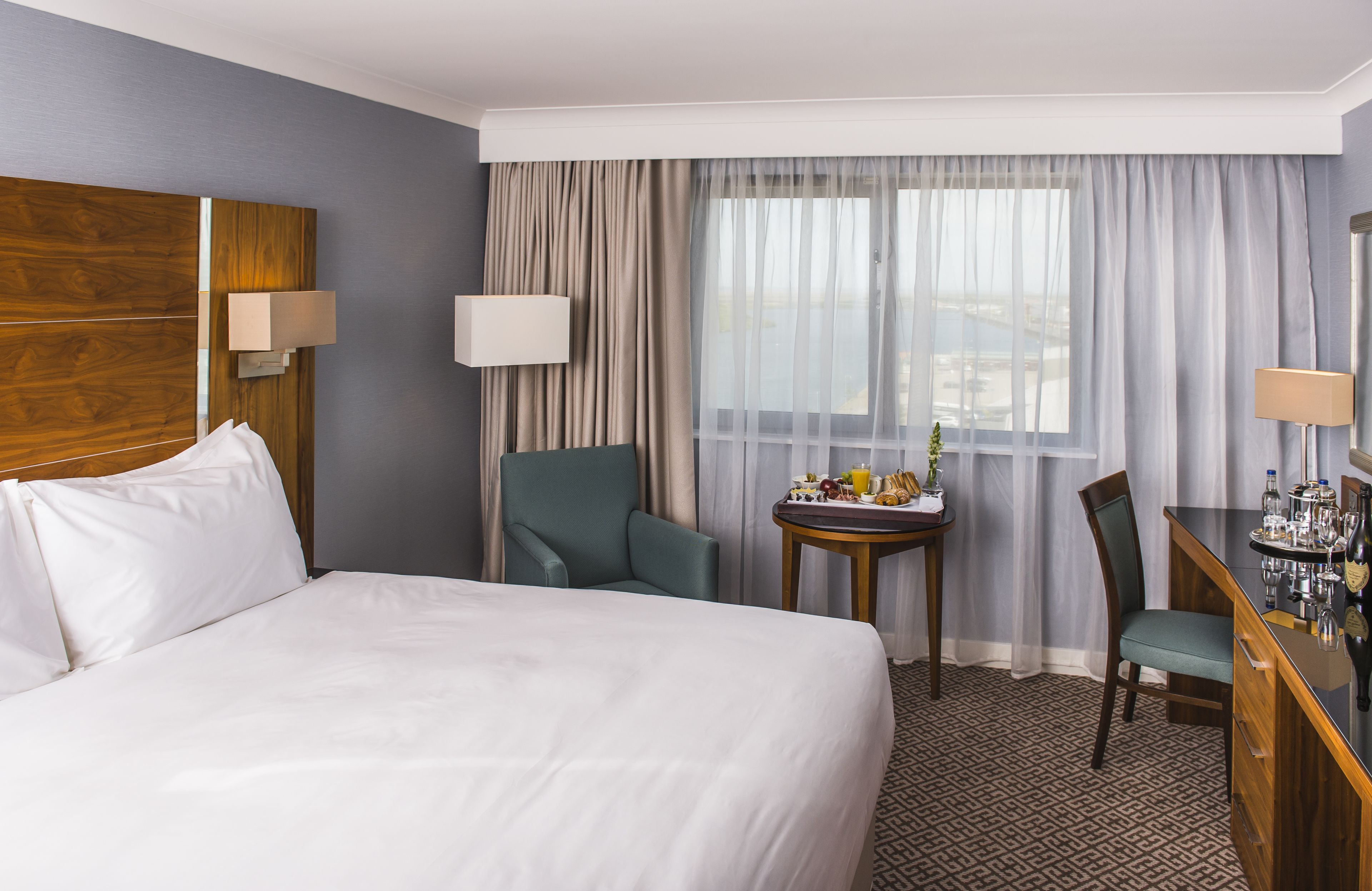 A hotel room with a large bed, a desk with a chair, a small table with breakfast items, and a view of the city through sheer curtains.
