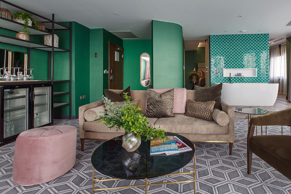 Ein modernes Wohnzimmer mit einer grünen Akzentwand, einem beigen Sofa, einem Glastisch und einem rosa Ottomanen.