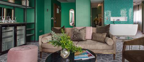 Un salon moderne avec un mur d’accent vert, un canapé beige, une table basse en verre et un pouf rose.