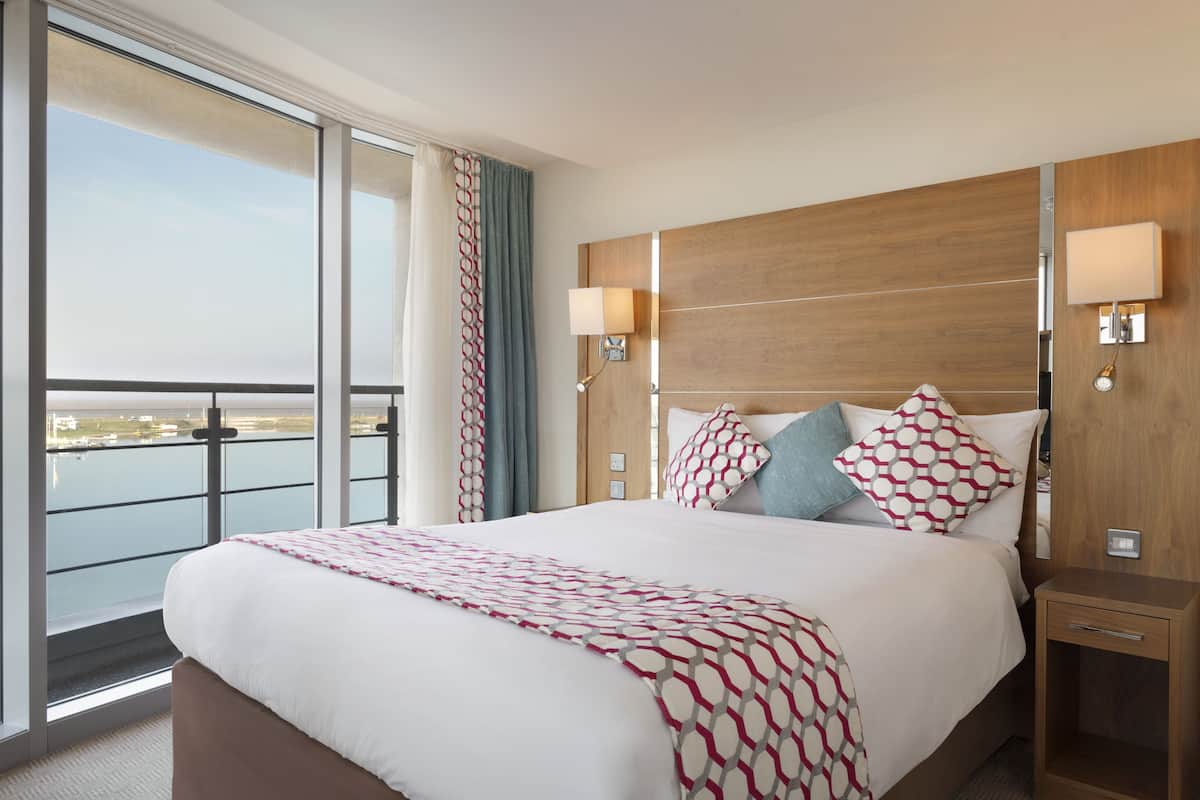 Ein Hotelzimmer mit einem großen Bett, zwei Nachttischlampen, einem hölzernen Kopfteil und Blick auf das Wasser durch große Fenster.