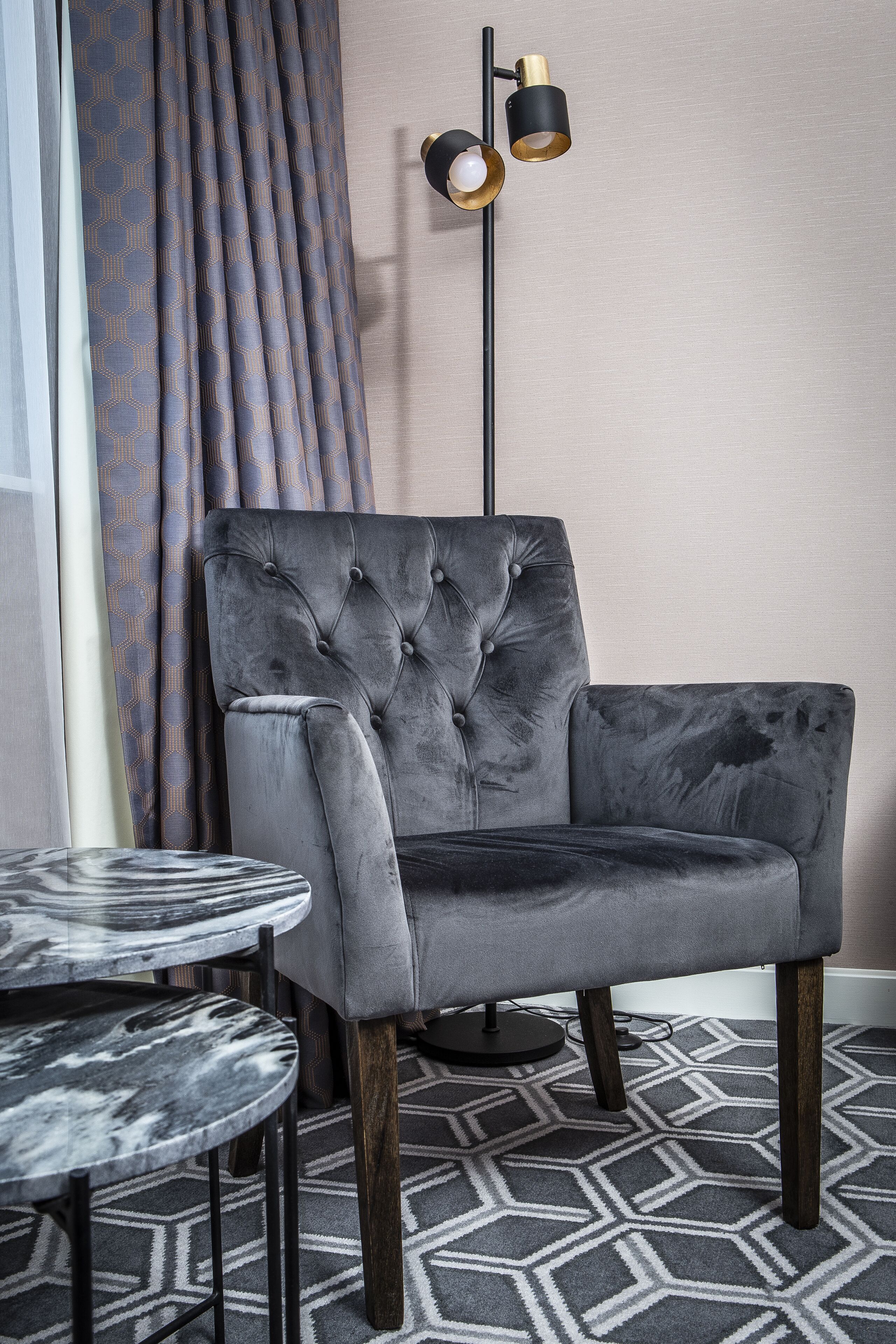 A grey upholstered armchair with a tufted backrest, a round marble table, and a patterned floor rug.