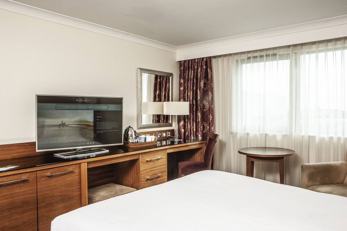 Ein Hotelzimmer mit Bett, Schreibtisch mit Fernseher, Sessel, kleinem Tisch und Vorhängen.