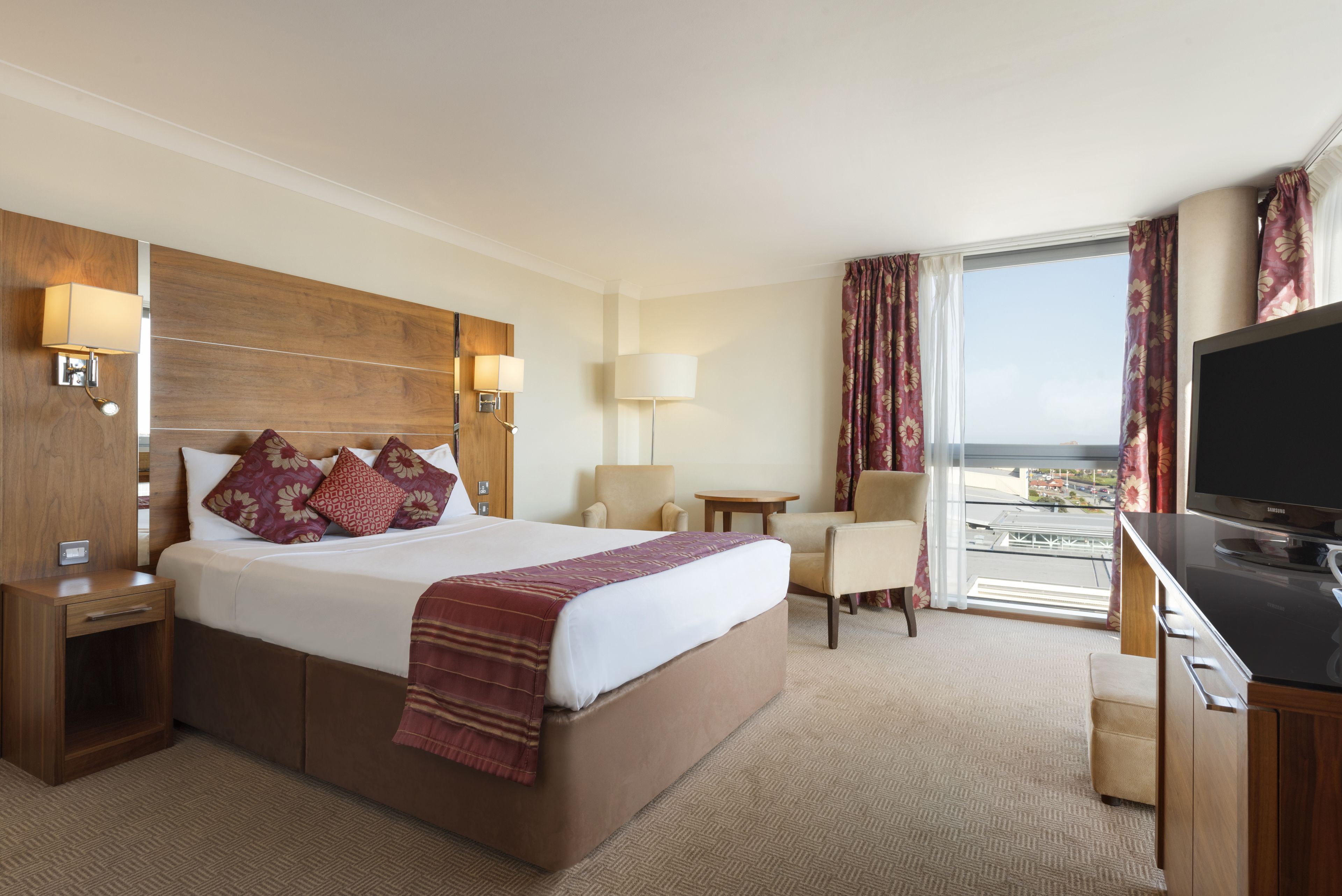 A hotel room with a large bed, a desk, a TV, and a view of the city.