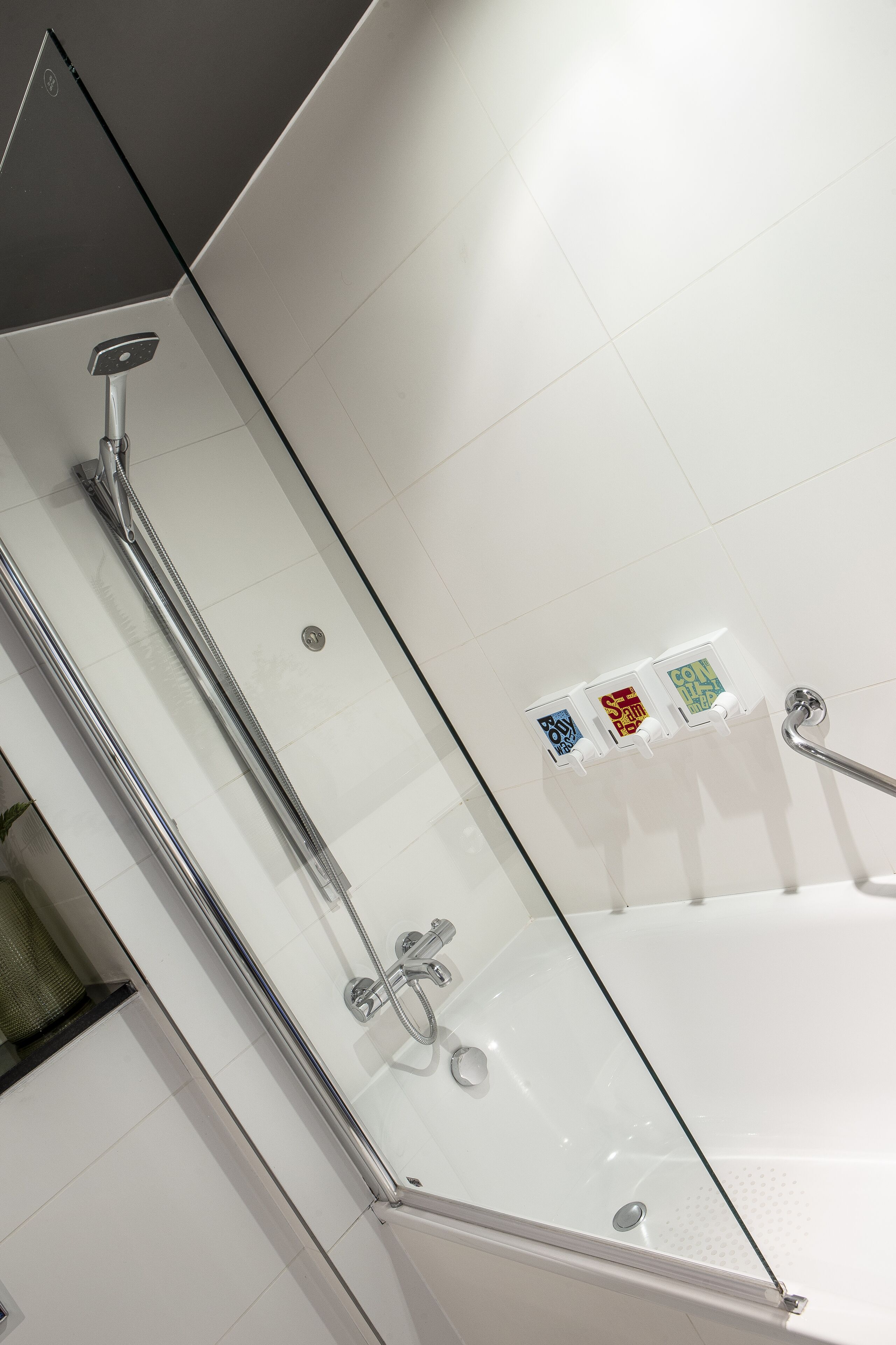 Un baño moderno con ducha envuelta en vidrio, una bañera y tres dispensadores de jabón de colores en la pared.
