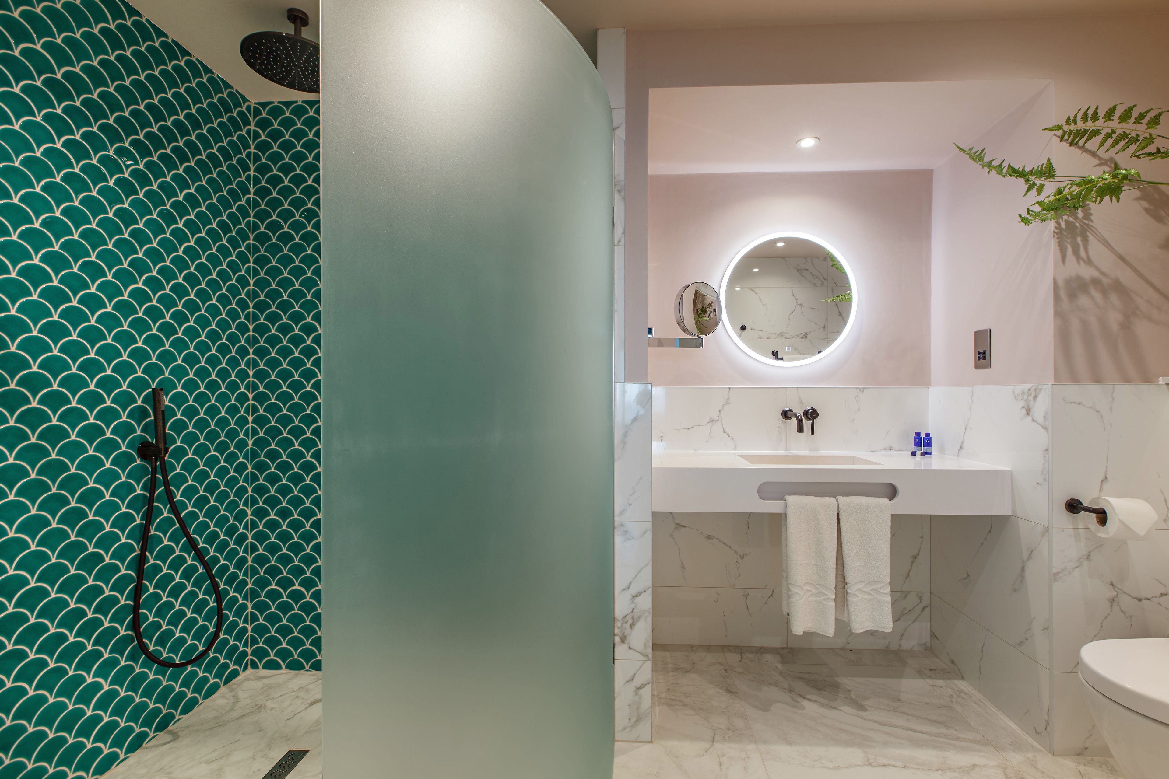 A bathroom with a teal tiled wall, a white sink with a marble countertop, and a round mirror with a light.