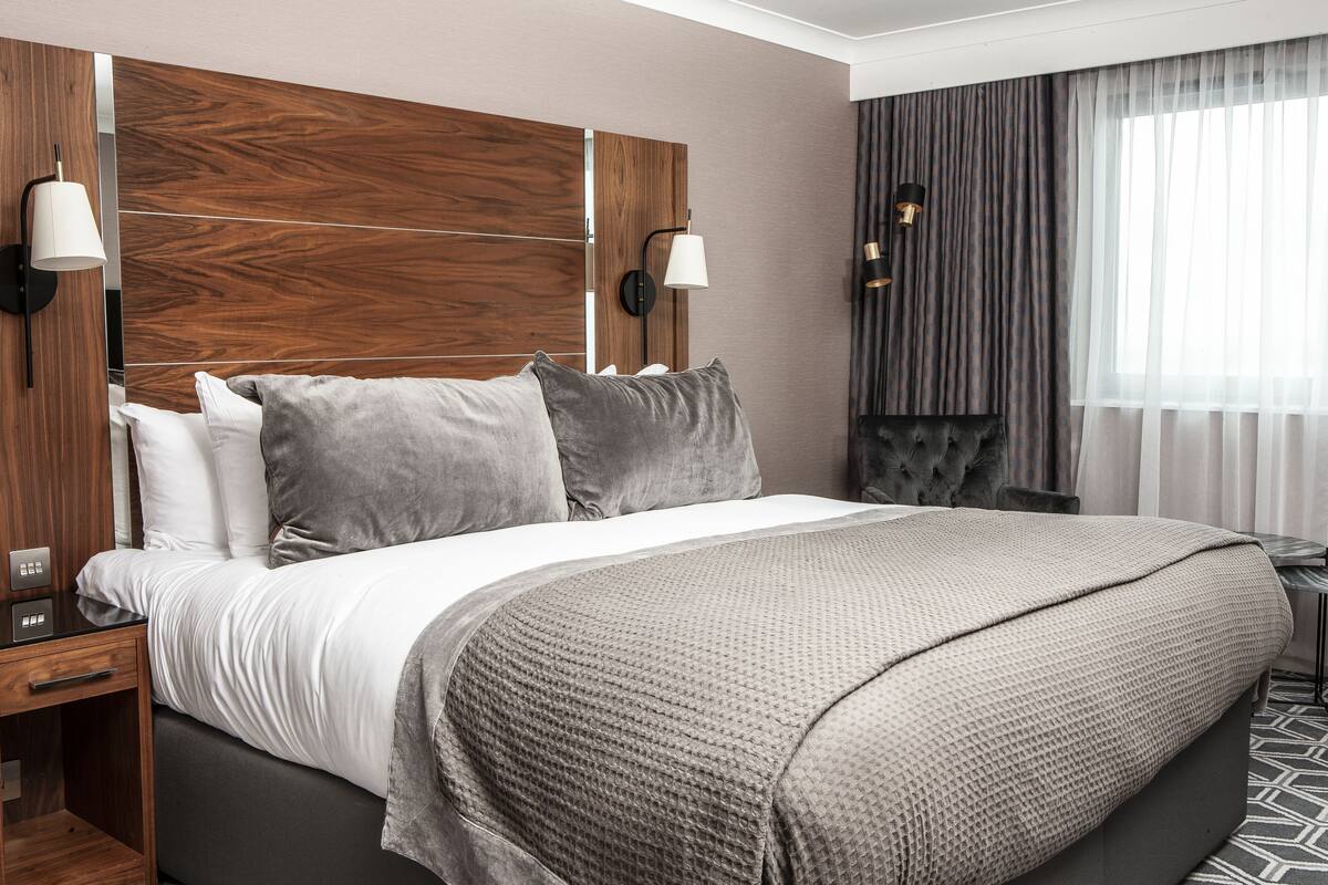 Ein modernes Hotelzimmer mit einem großen Bett, einem Holzkopfende, zwei Nachttischlampen, einem kleinen Tisch und Blick ins Freie durch transparente Vorhänge.