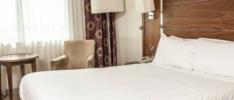 A hotel room with a large bed, a chair, a small table, a lamp, and a telephone.