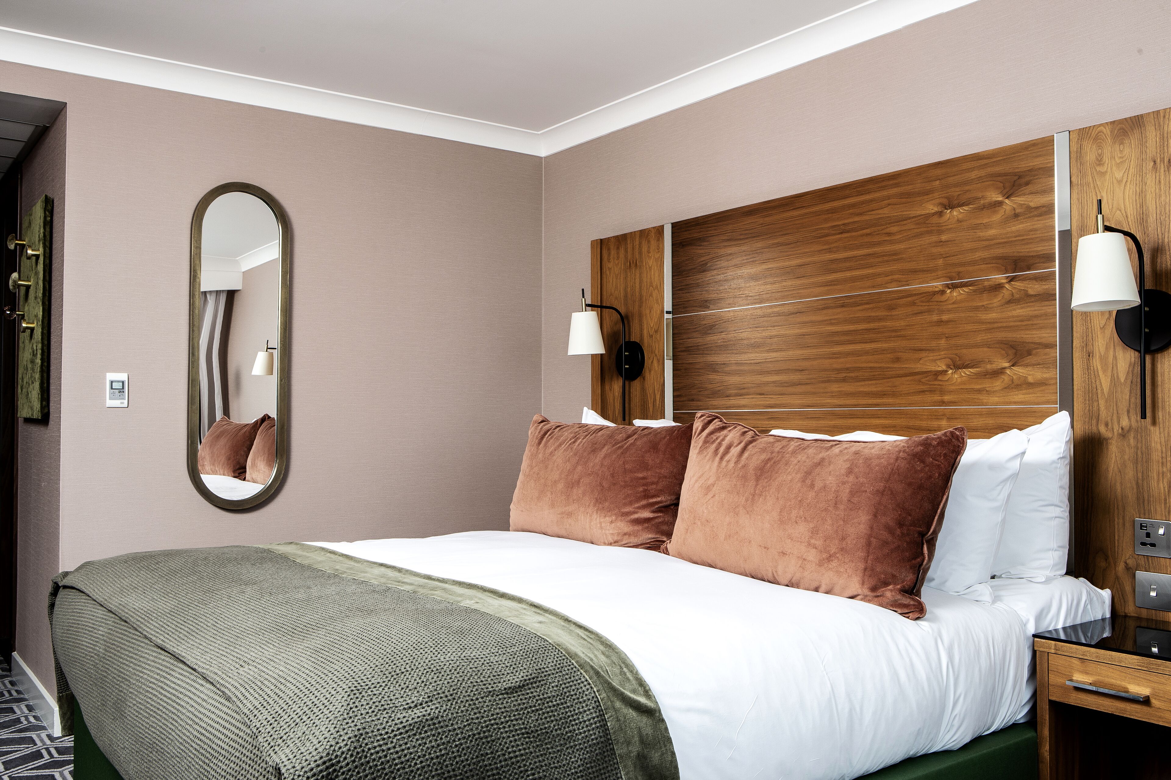 A hotel room with a large bed, two wall-mounted lamps, a wooden headboard, and a mirror.