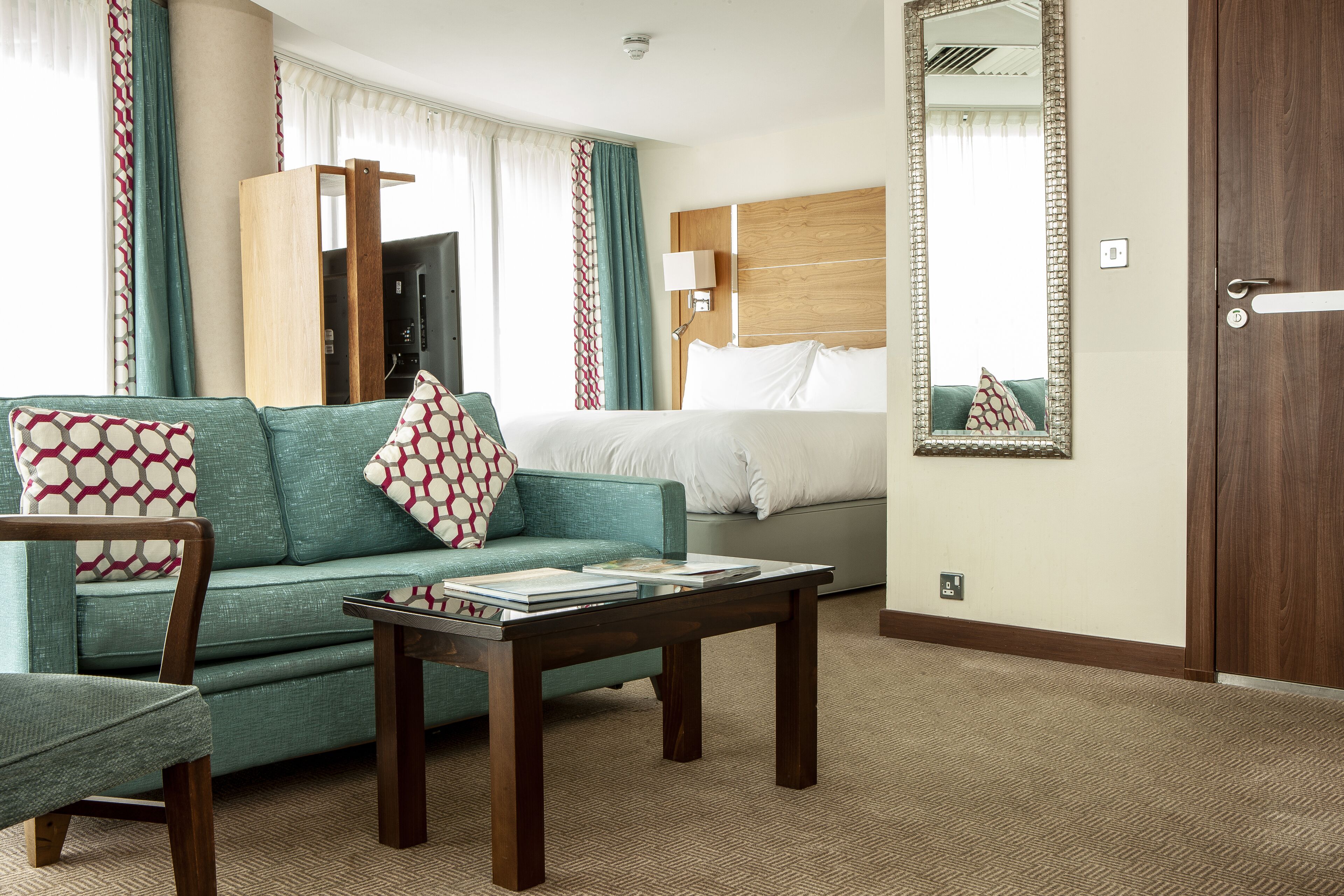 A hotel room with a sofa, a chair, a small table, a bed, a mirror, and a door.
