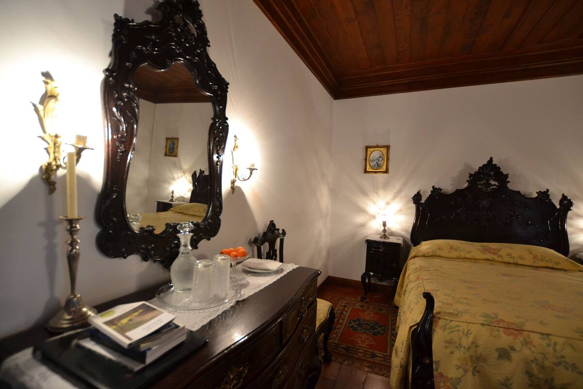 A bedroom with a wooden bed, a large ornate mirror, a dresser with books, and a nightstand with a lamp.