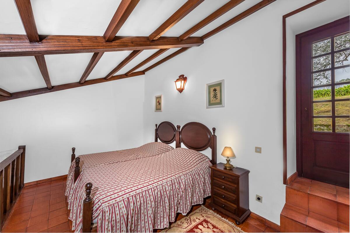 A bedroom with a wooden bed, a nightstand, a wall-mounted lamp, and a door with a view of greenery.