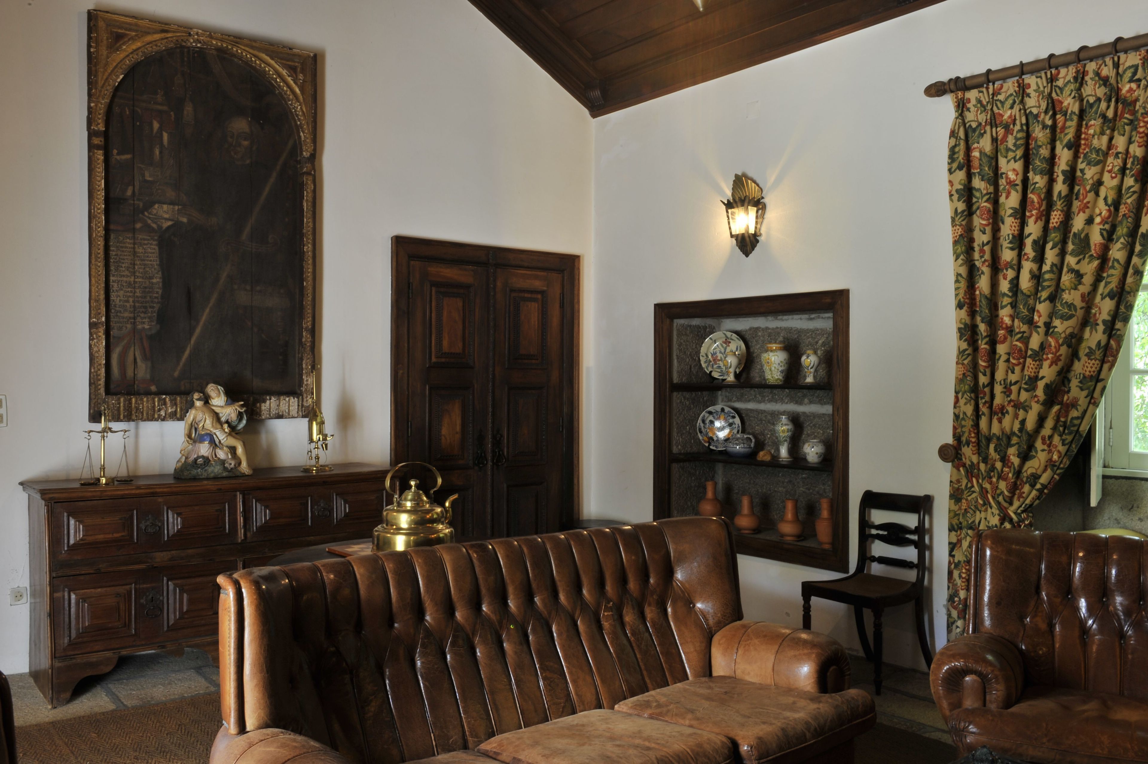Una habitación con un armario de madera, un sofá, una silla, un estante montado en la pared con objetos decorativos y un cuadro grande enmarco.