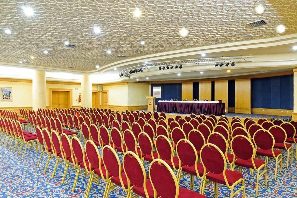 A conference room with rows of red chairs, a stage with a purple backdrop, and a carpeted floor.