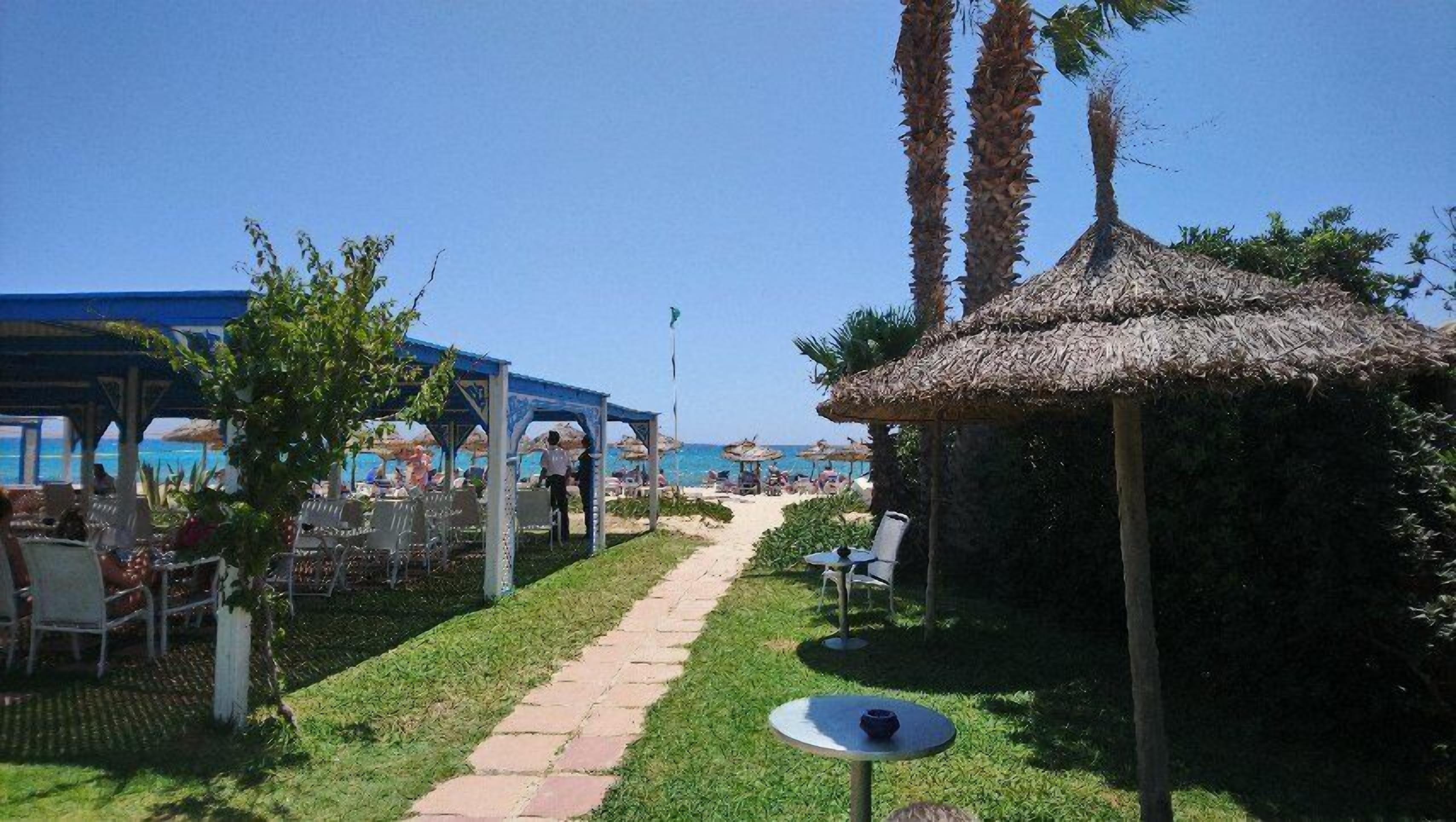 Un resort frente a la playa con mesas al aire libre, una estructura de techo de paja y vista despejada al océano.