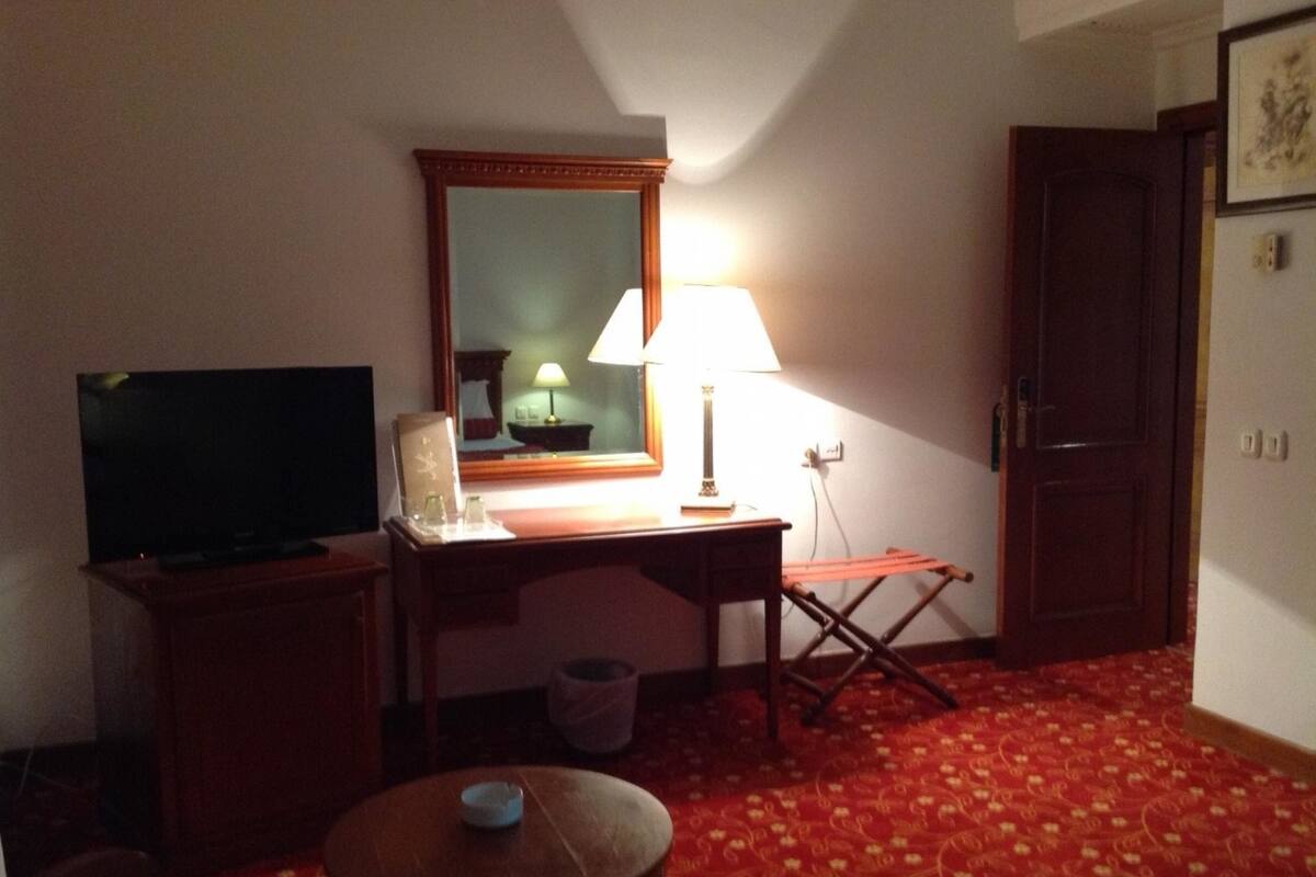 A hotel room with a television, a desk with a lamp, a small table, and a red patterned carpet.