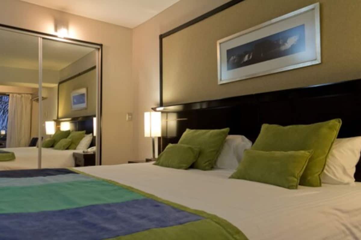 Une chambre d’hôtel avec un grand lit, des oreillers verts, une tête de lit, un tableau encadré au mur et un miroir qui reflète la pièce.