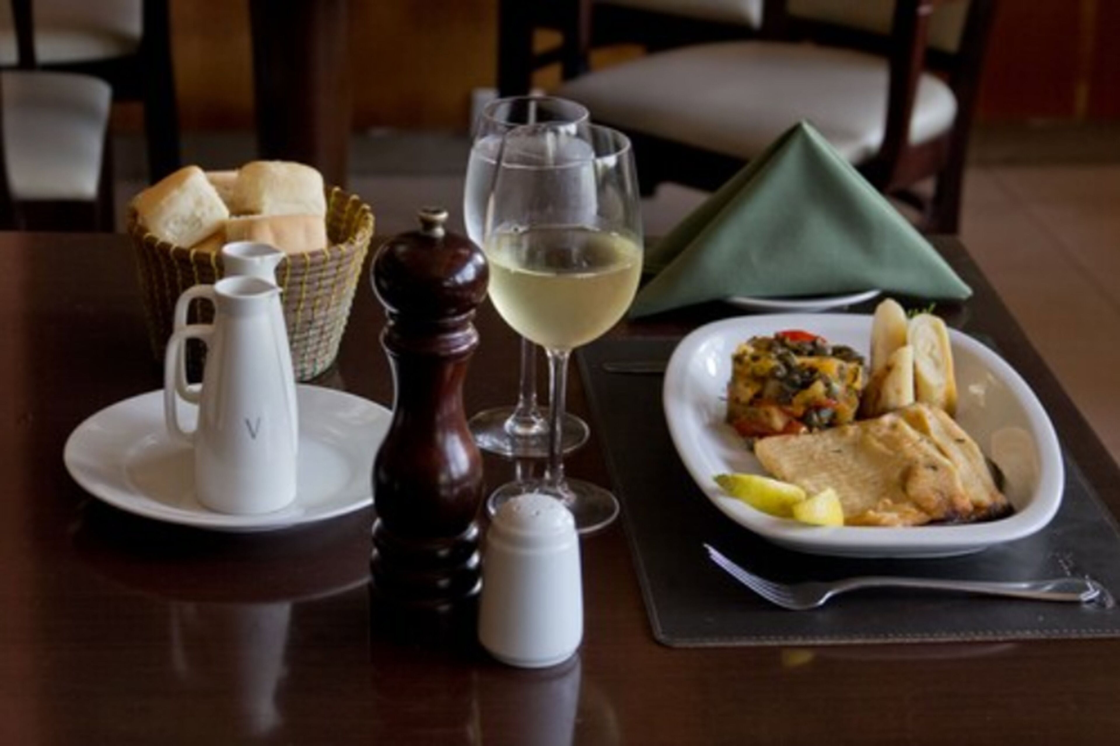 A dining table with a pitcher, salt and pepper shakers, a basket of bread, a glass of white wine, and a plate of food.