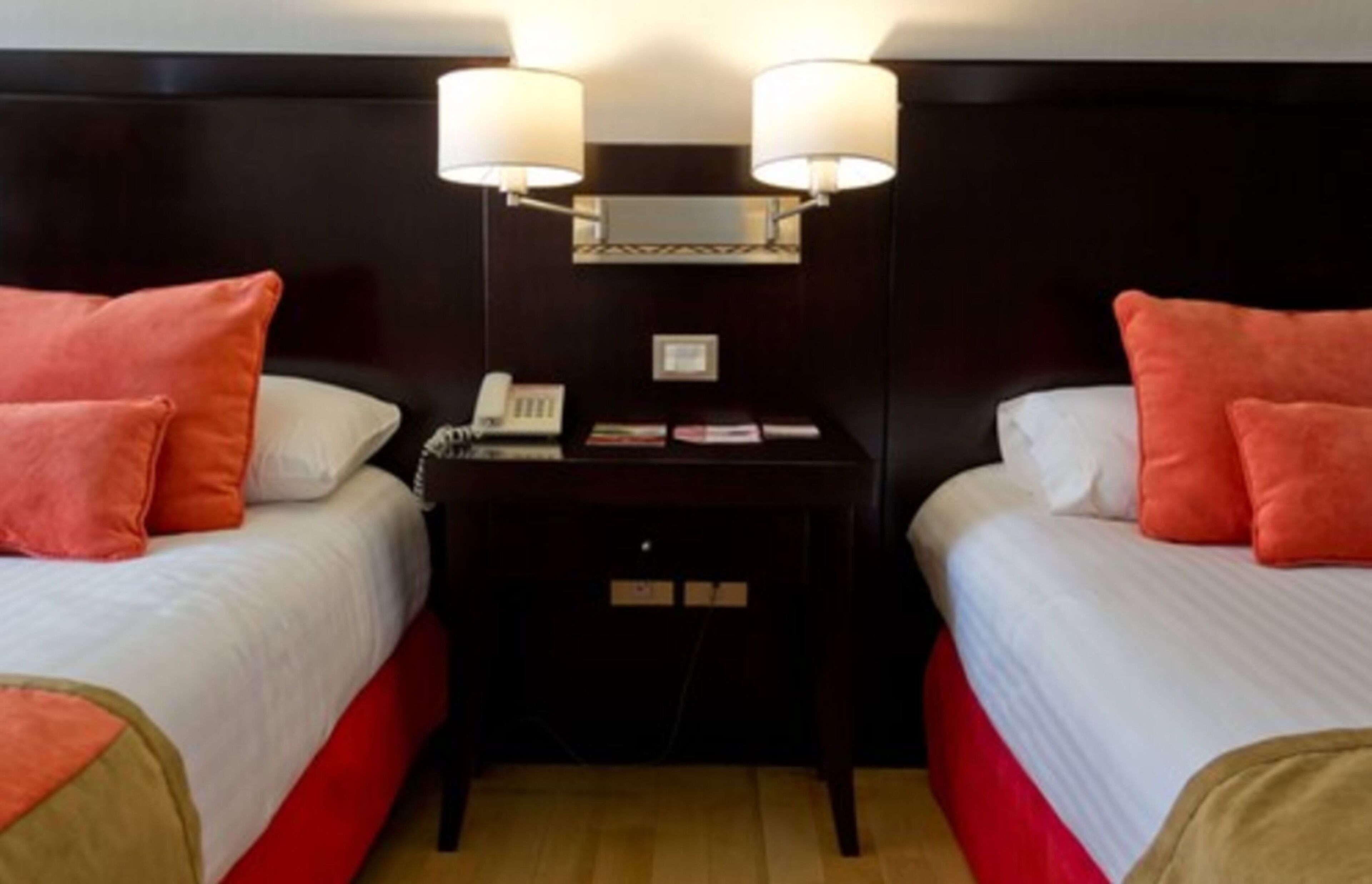 Ett hotellrum med två sängar, båda med vita och röda sängkläder, ett litet bord med en telefon och tidskrifter samt två väggmonterade lampor.