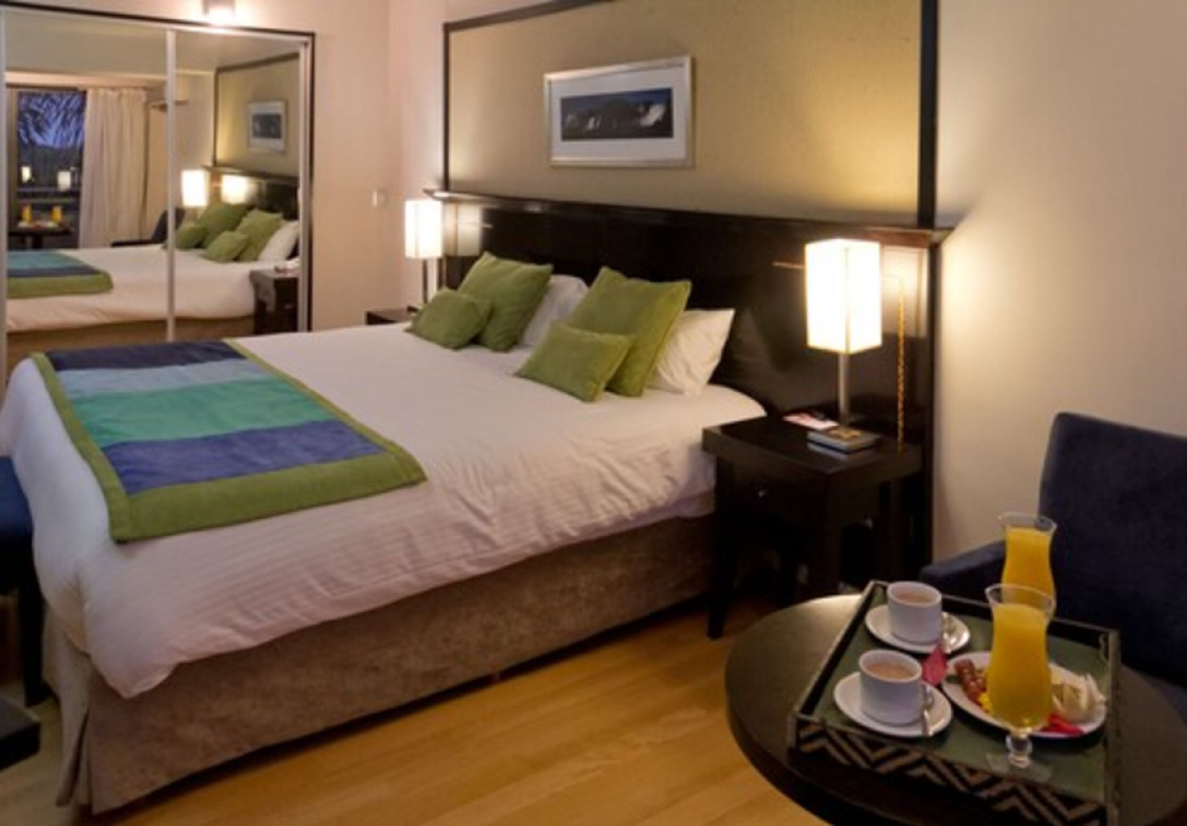 A hotel room with a large bed, a bedside table with a lamp, a chair, and a tray with breakfast items.