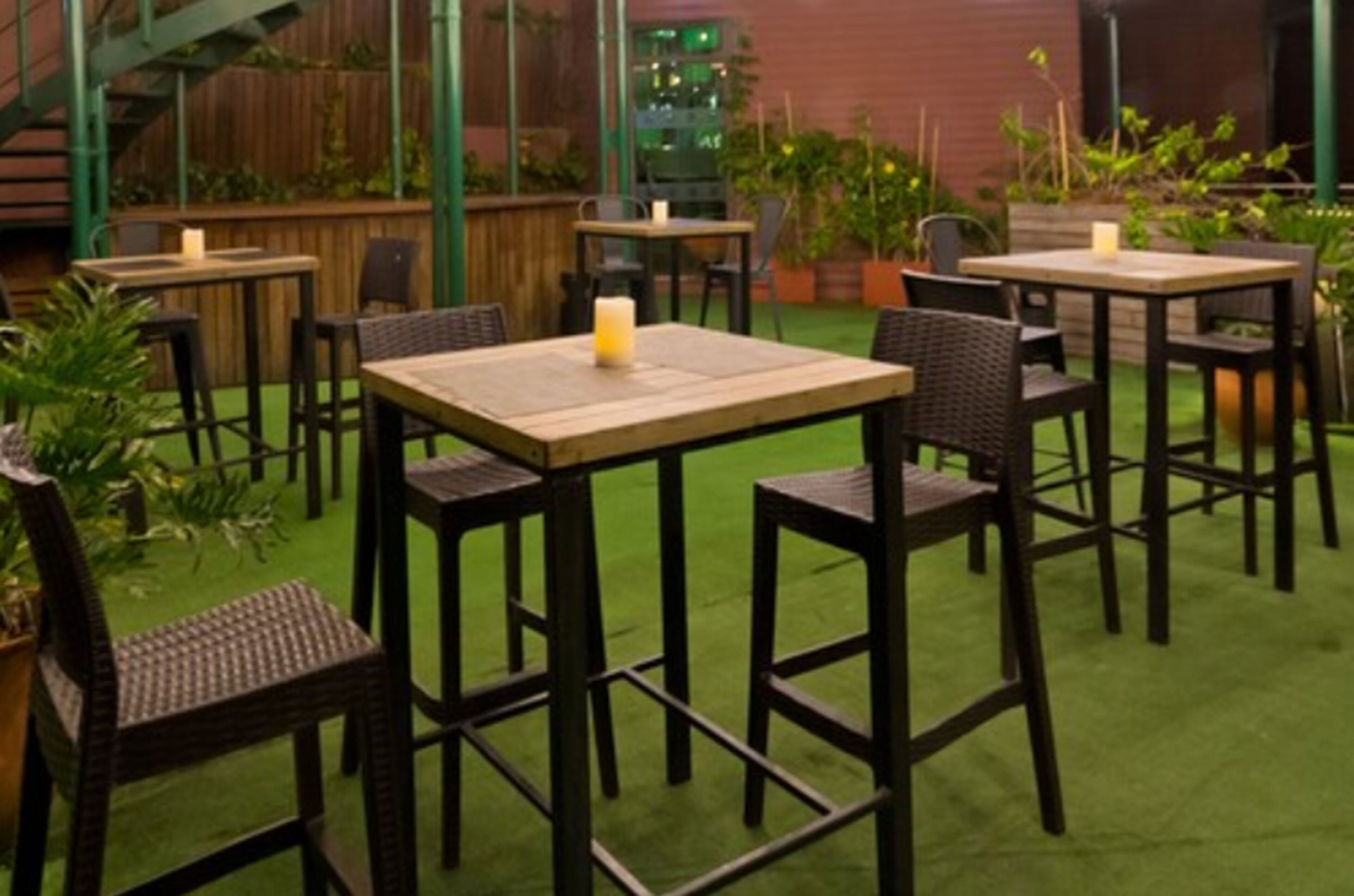 An outdoor patio area with tables and chairs, surrounded by greenery and a wooden fence.