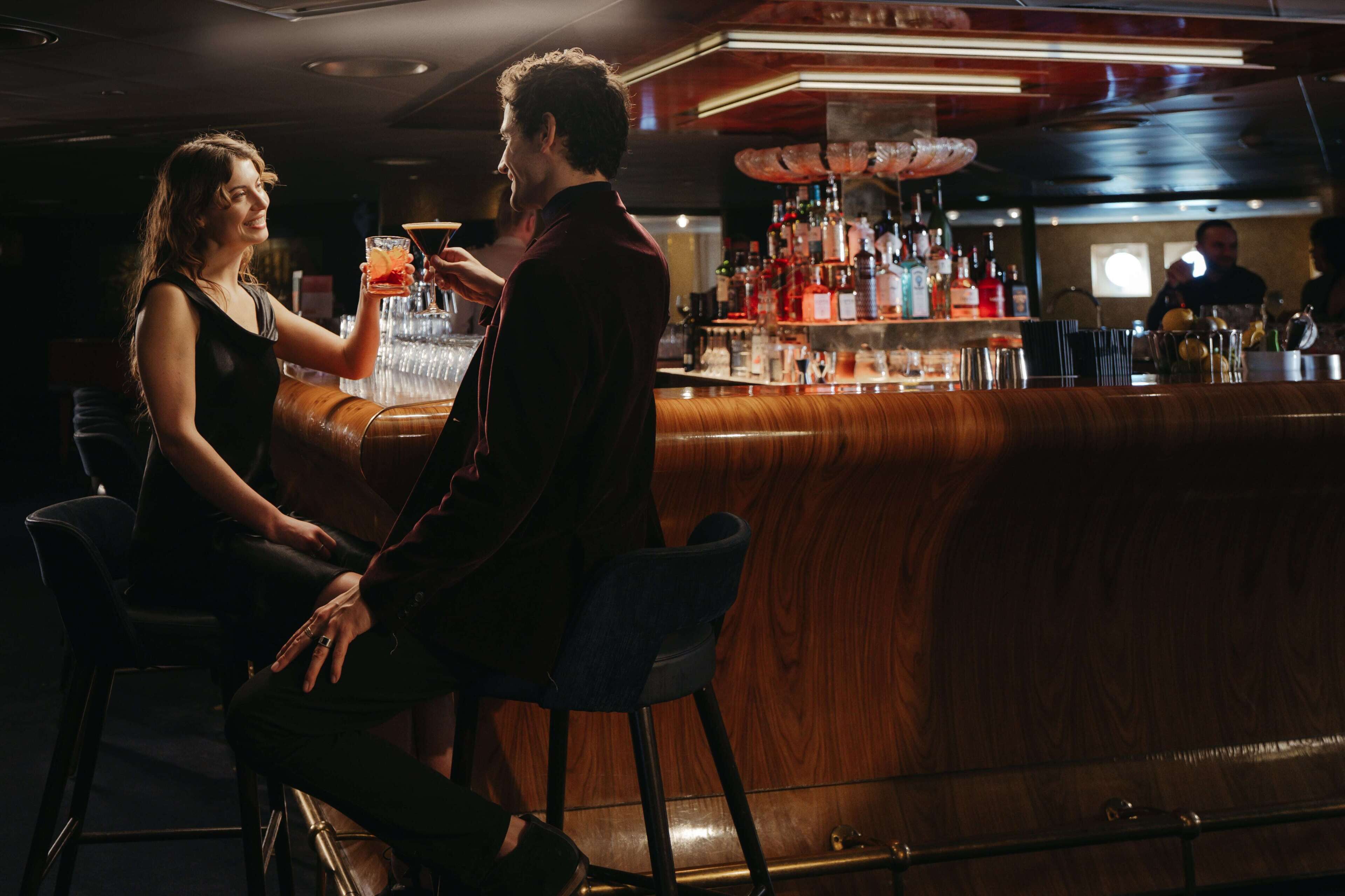 A bar area with a wooden counter and stools, a bar stocked with bottles, and a person holding a drink.