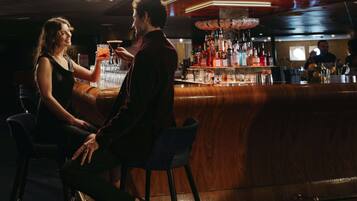 A bar area with a wooden counter and stools, a bar stocked with bottles, and a person holding a drink.