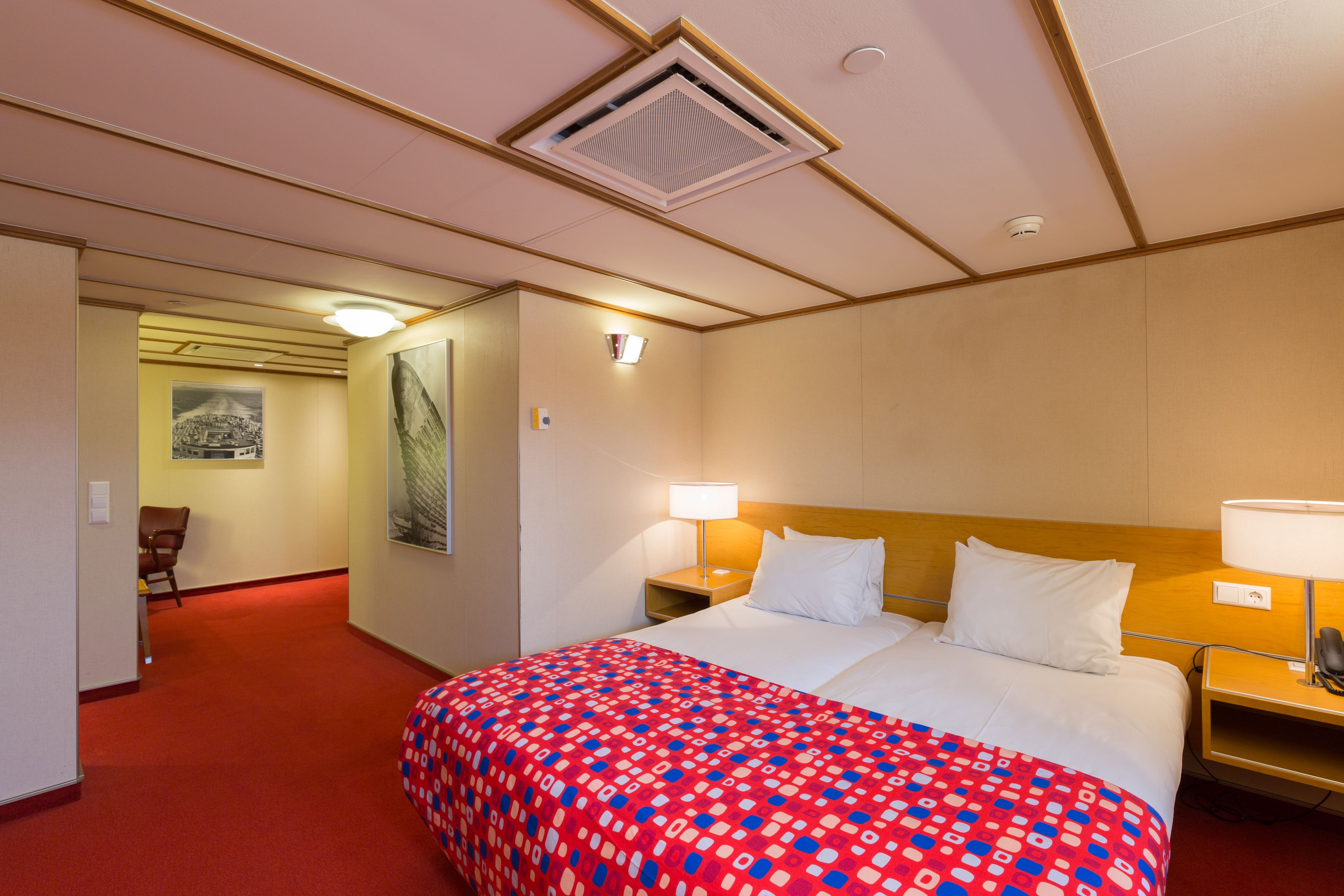 A hotel room with a large bed, two bedside tables with lamps, a red carpeted floor, a wooden headboard, and a framed picture on the wall.