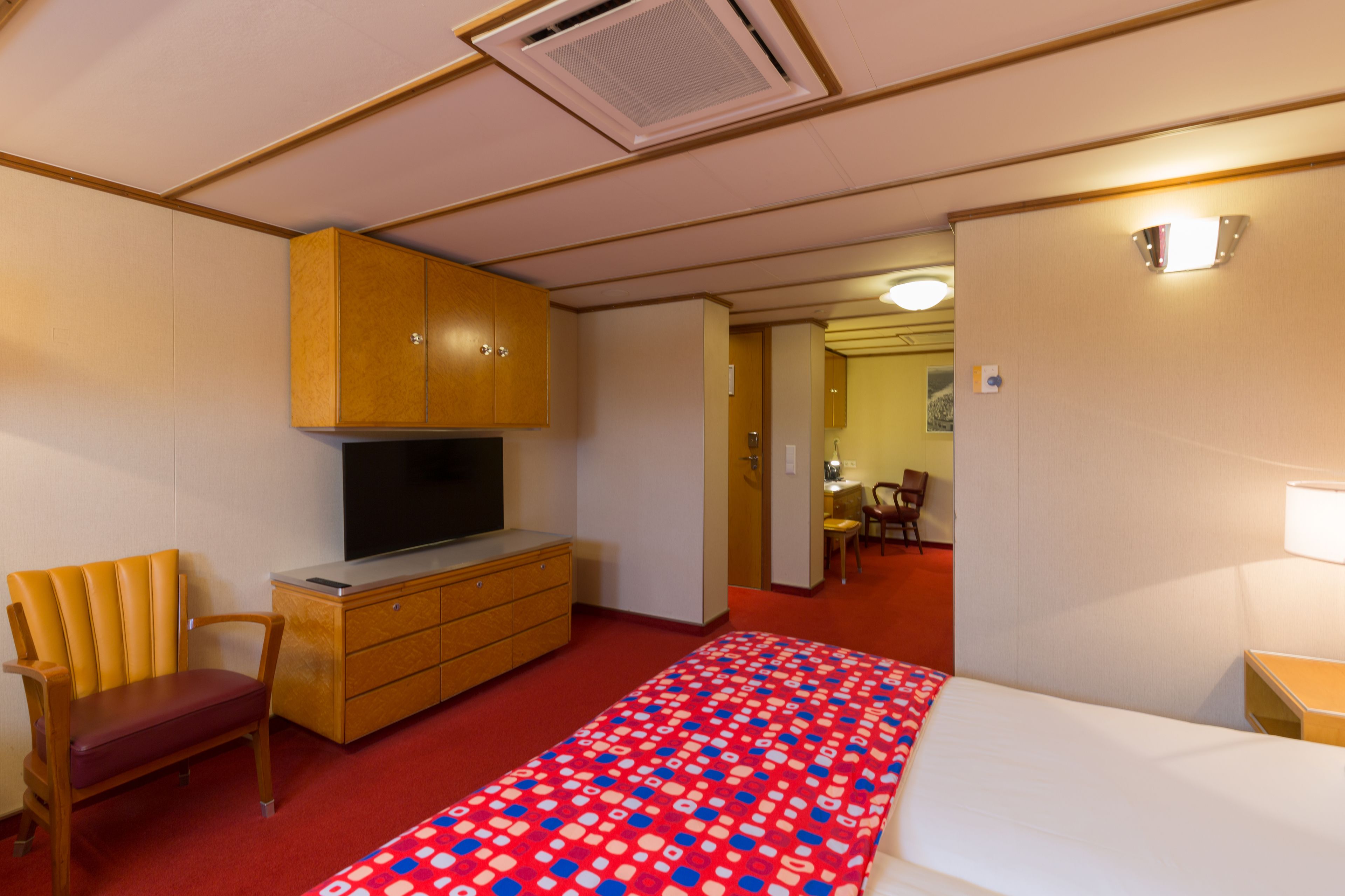 Una camera d'albergo con un letto, una televisione, un armadio in legno, una sedia e una moquette rossa.
