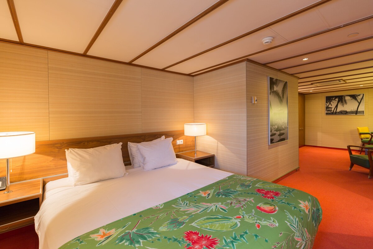 A hotel room with a bed, two bedside lamps, a nightstand, a wooden headboard, a wall-mounted picture, and a small table with chairs.