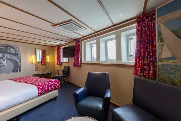 Une chambre d’hôtel comprenant un lit, deux chaises, un bureau, une télévision et de grandes fenêtres avec des rideaux.