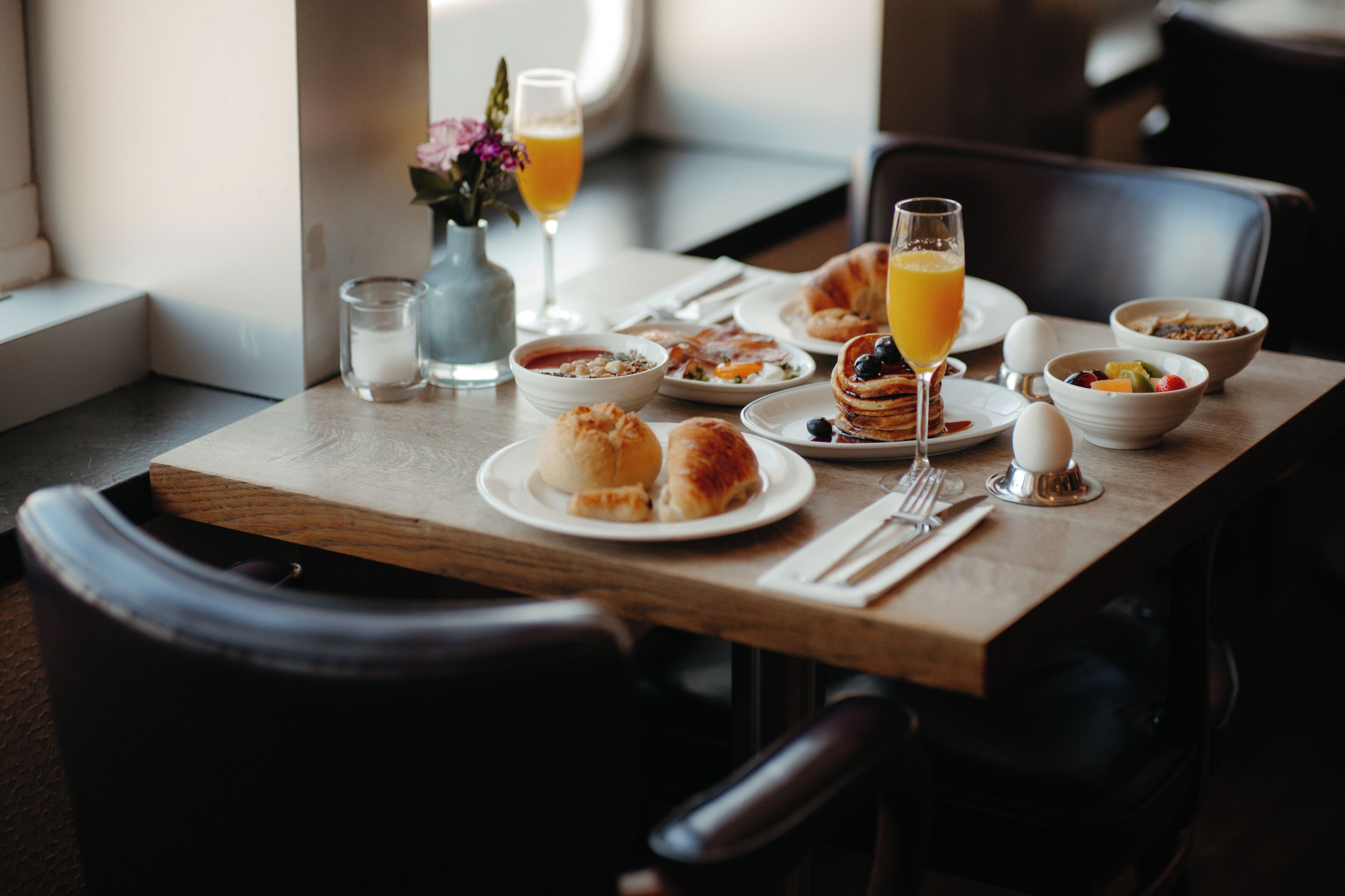 Una mesa de desayuno con platos de comida, un vaso de jugo de naranja y un florero con flores.