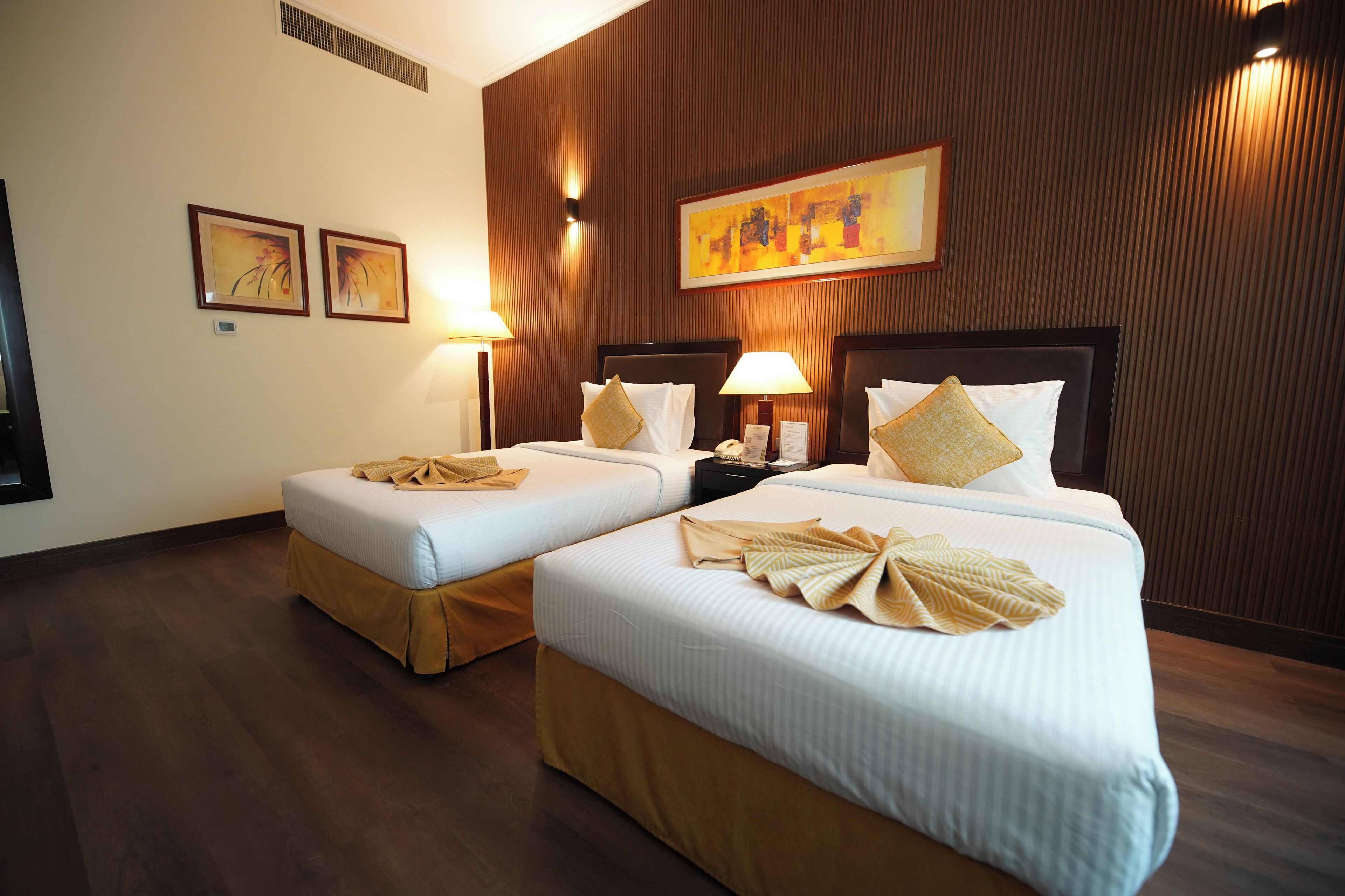 A hotel room with two beds, each with a pillow and a blanket. There are wall-mounted lamps and framed artwork on the wall.