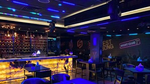 Een bar met een marmeren aanrecht, verschillende zithoeken en stoelen. Op de muur hangen neonborden, waaronder een met de tekst 'NIGHT CLUB'.
