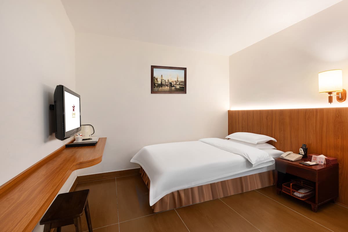 A hotel room with a bed, a desk with a computer, a nightstand, a wall-mounted TV, and a framed picture.