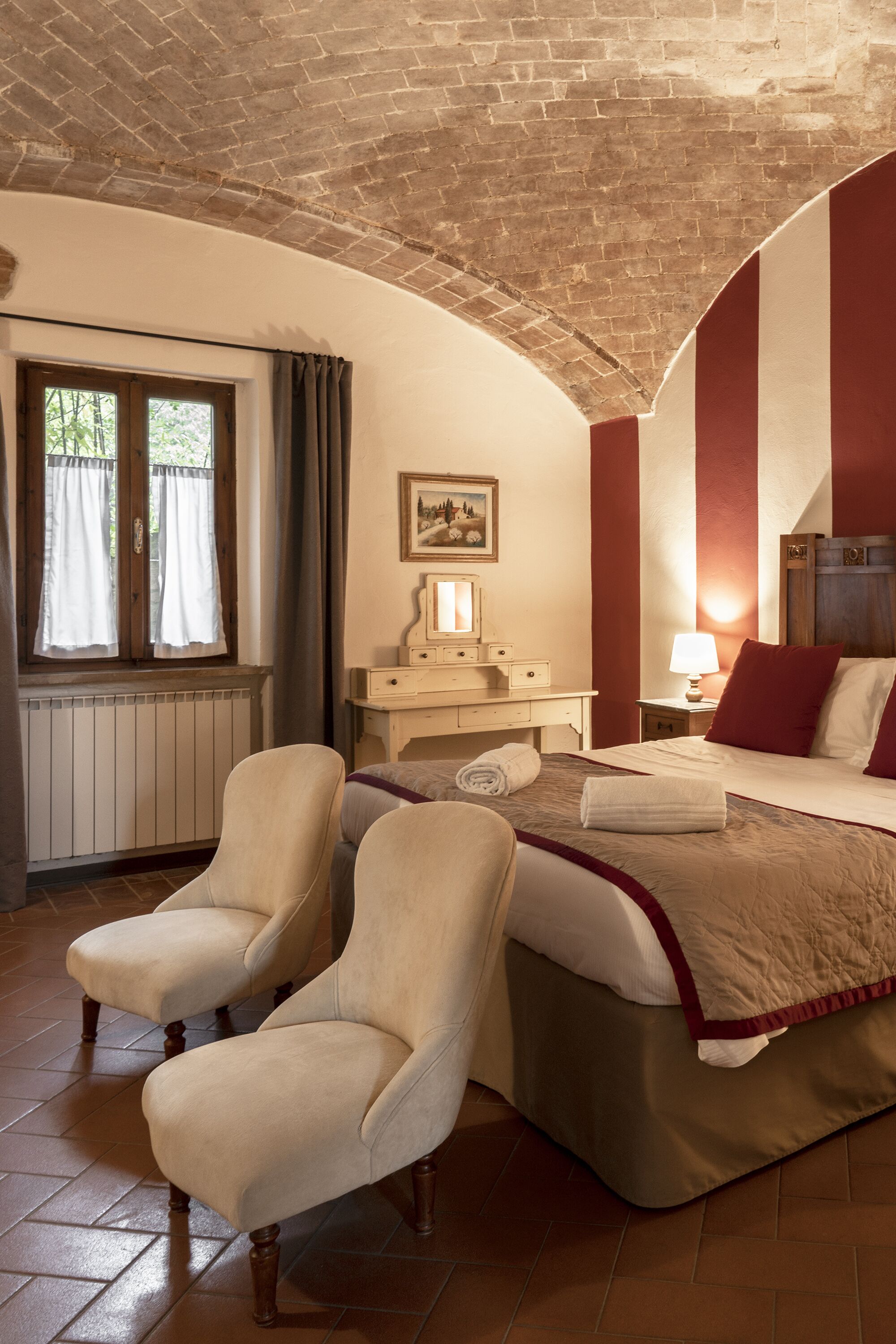 Un dormitorio con techo de ladrillo, una cama con ropa de cama roja y blanca, dos sillones, una mesita de noche con lámpara y una ventana con cortinas.
