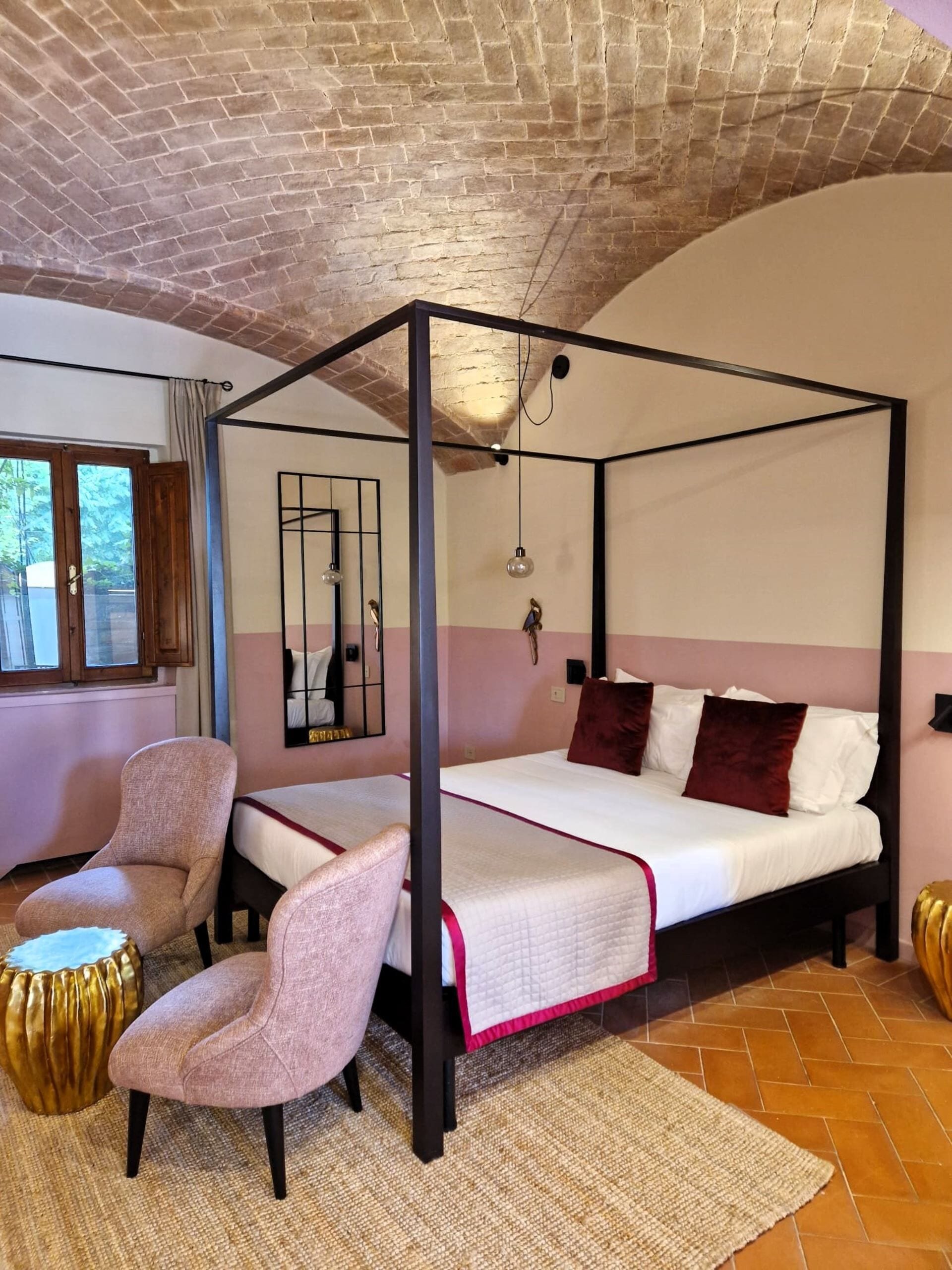A four-poster bed with a white and pink bedspread, two upholstered chairs, a gold accent chair, a woven rug, and a brick ceiling.