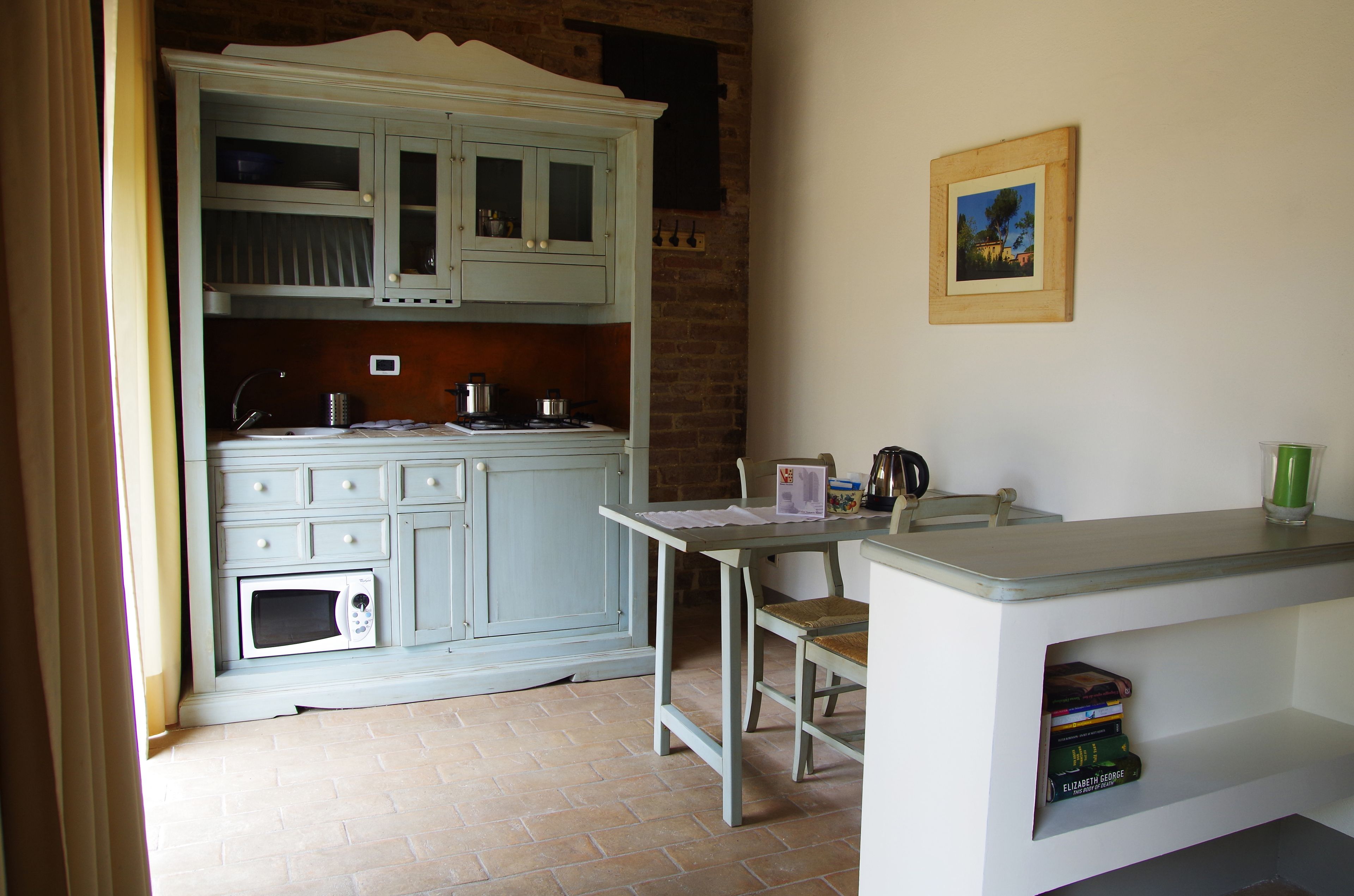 A kitchen with a microwave, stove, and cabinets. There is a small table with a chair and a shelf with books.