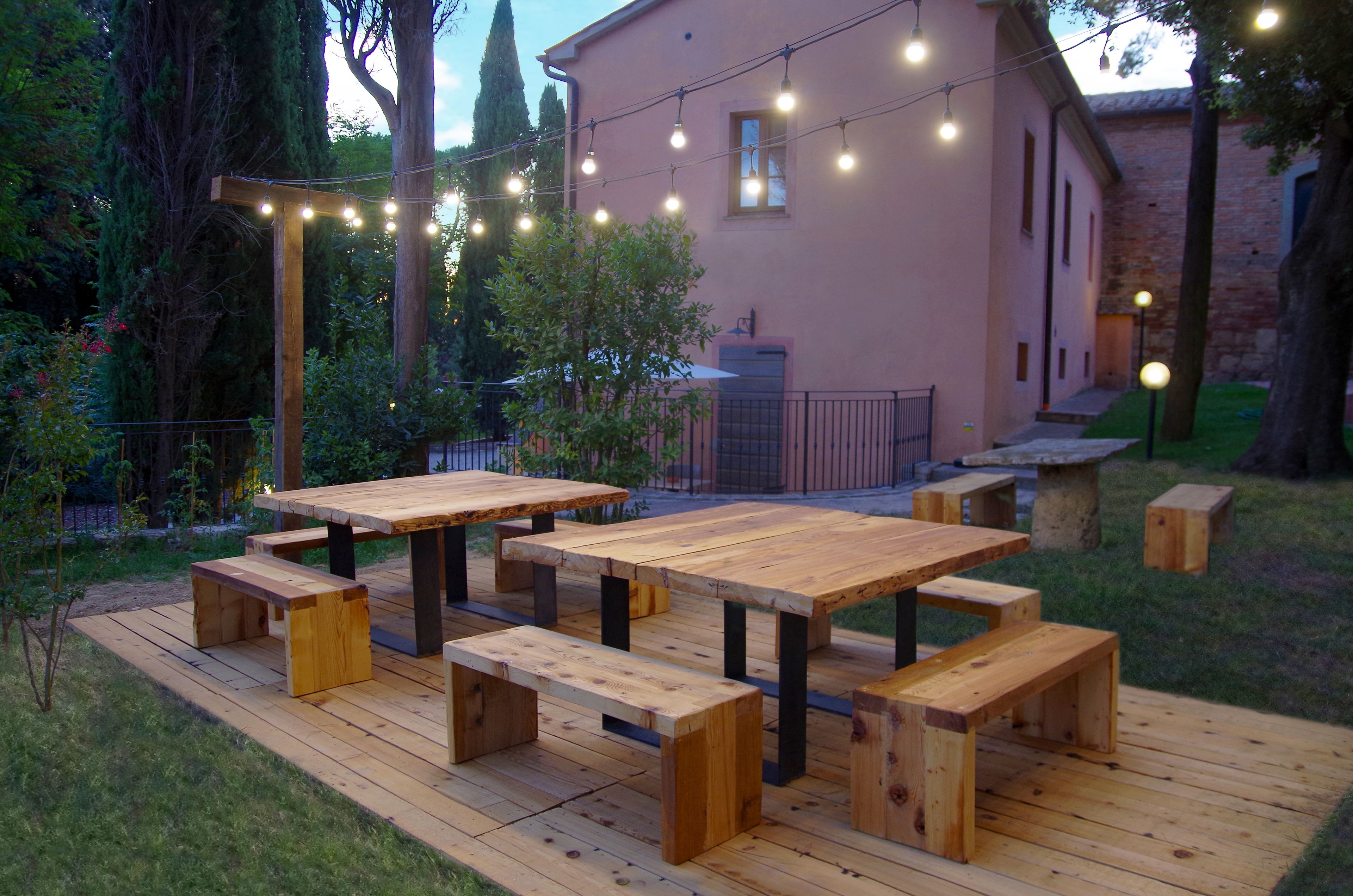 An outdoor wooden deck with picnic tables and benches, surrounded by greenery and a building in the background.