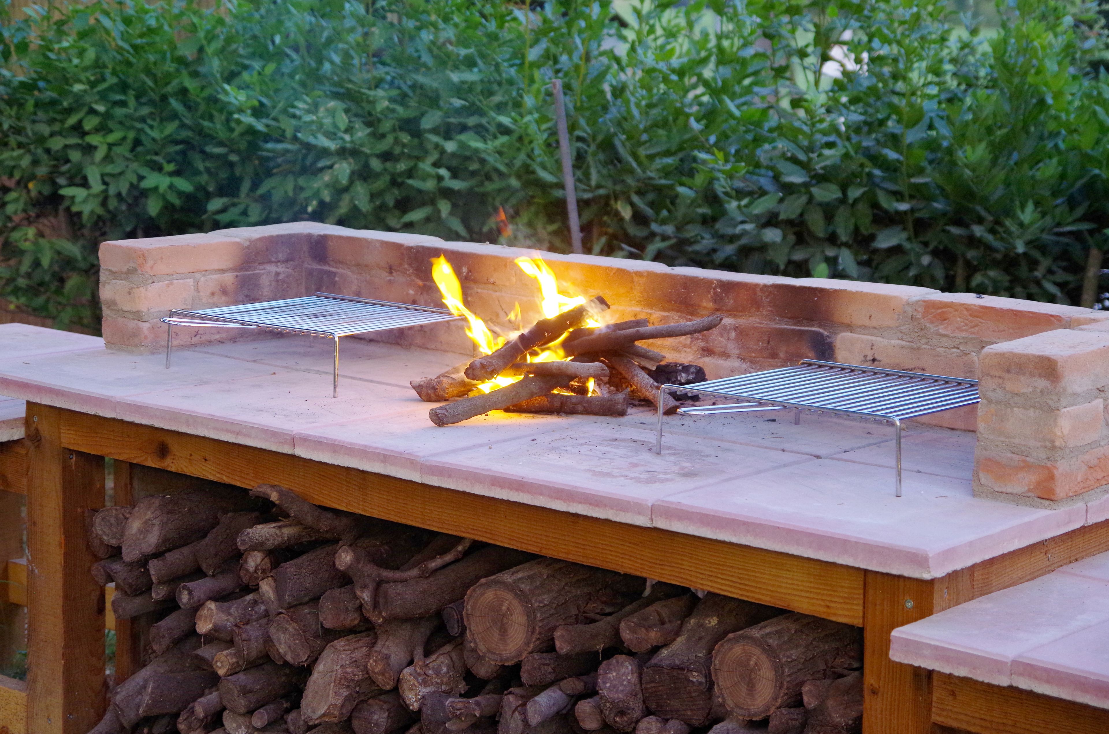 An outdoor grill with two grilling stations, surrounded by a brick wall and a stack of firewood.