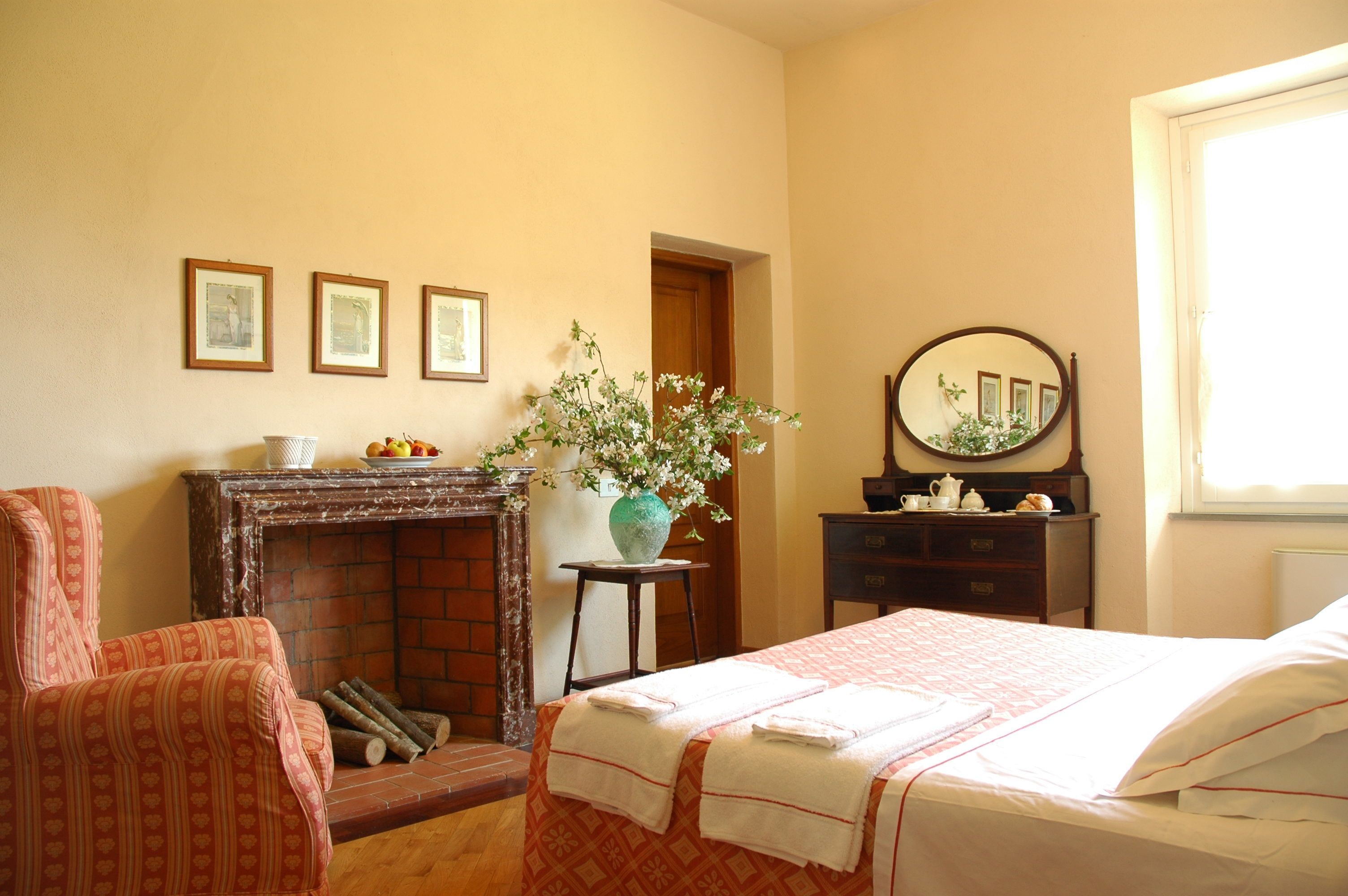 Quarto com lareira, cama com lençóis brancos, espelho e quadros nas paredes.