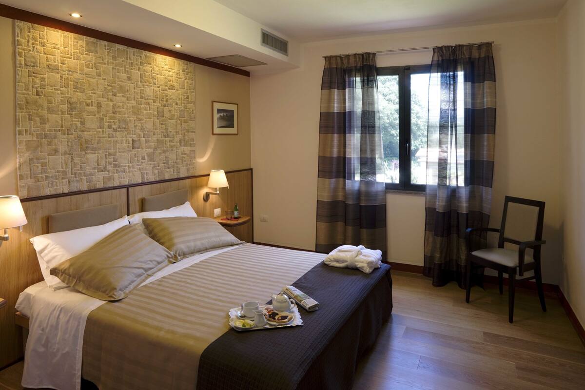 Una habitación de hotel con pared de piedra, una cama con cabecera de madera, una silla y un ventanal con cortinas.