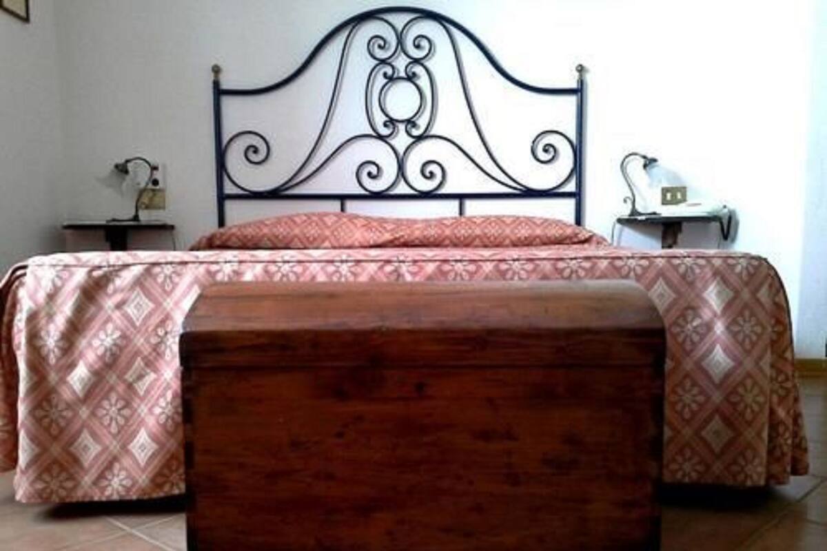 Una cama con una colcha estampada, un banco de madera, una mesita de noche con una lámpara y un estante fijado a la pared.