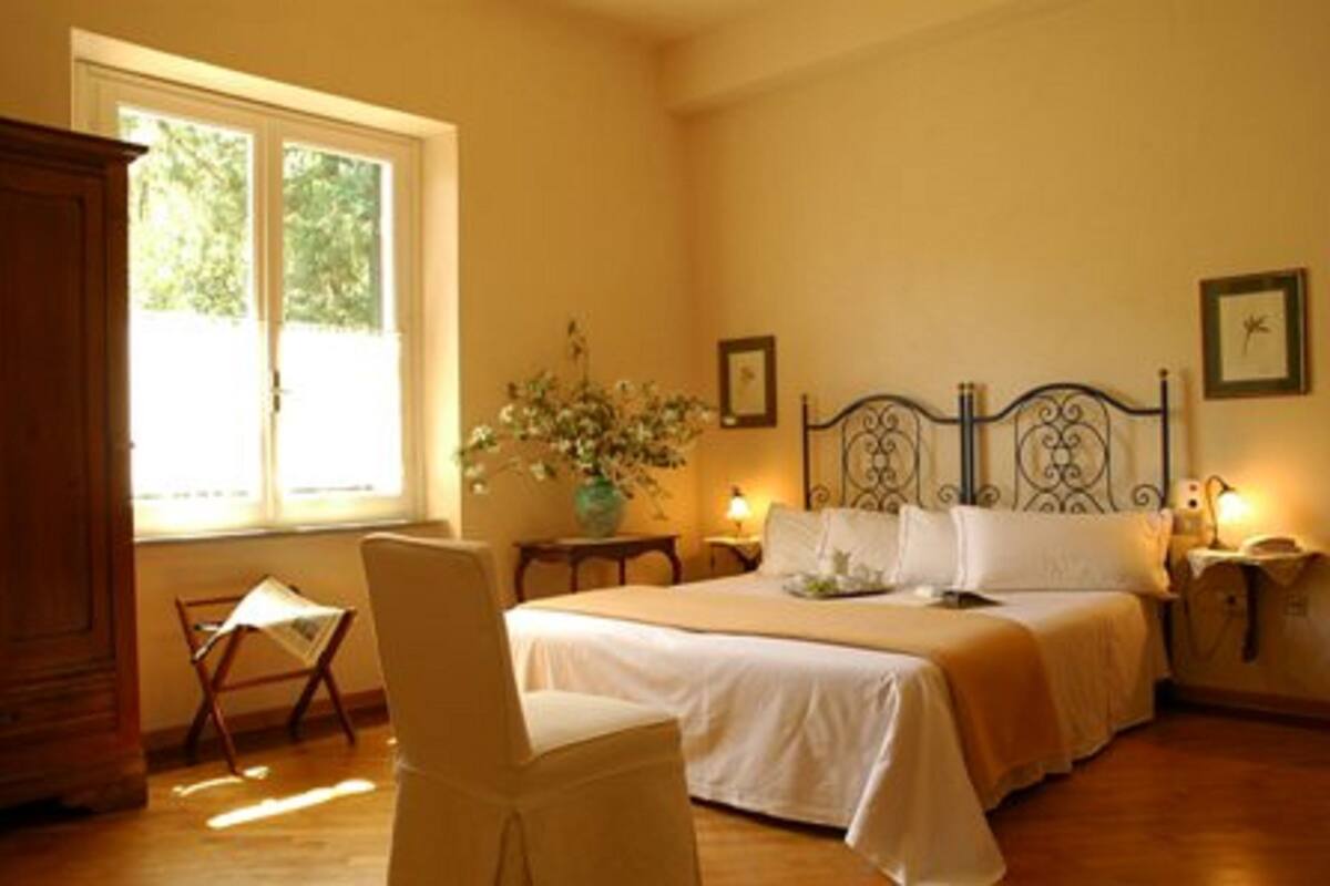 Un dormitorio con cama, una silla, un ventanal y una mesita de noche con lámpara.