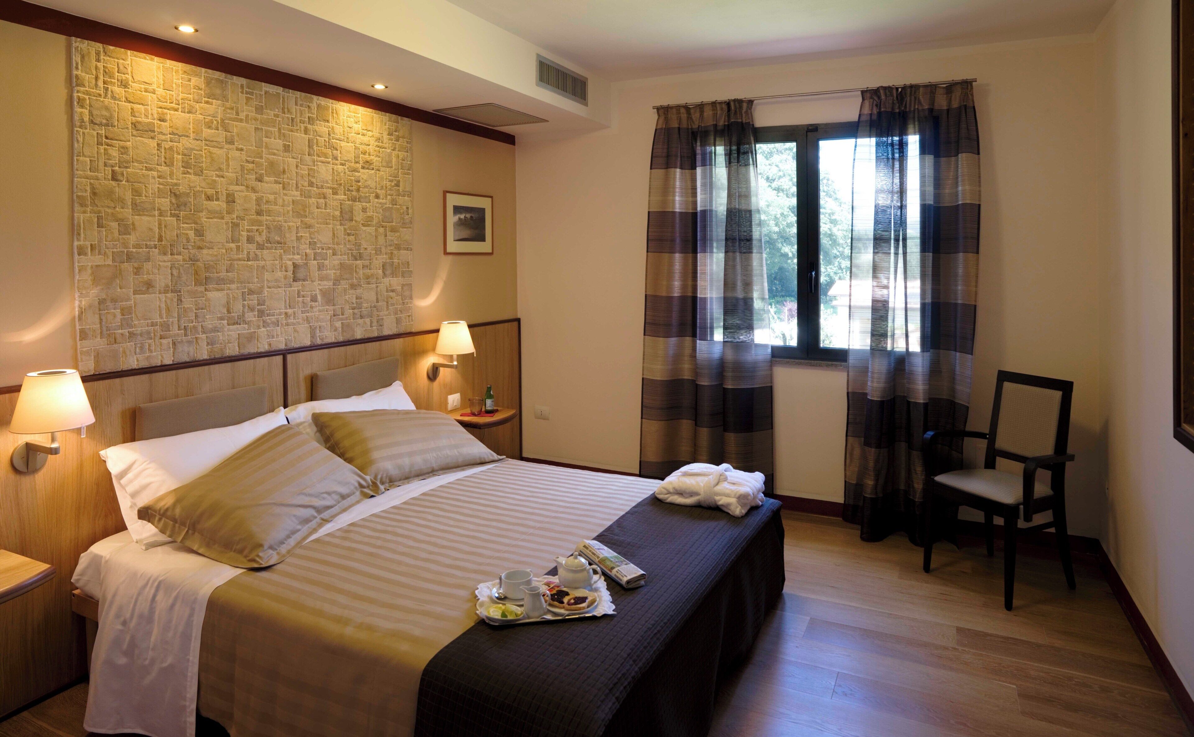 Habitación de hotel con pared de piedra, cama con cabecero, mesita de noche con lámpara, una silla y ventana con cortinas.