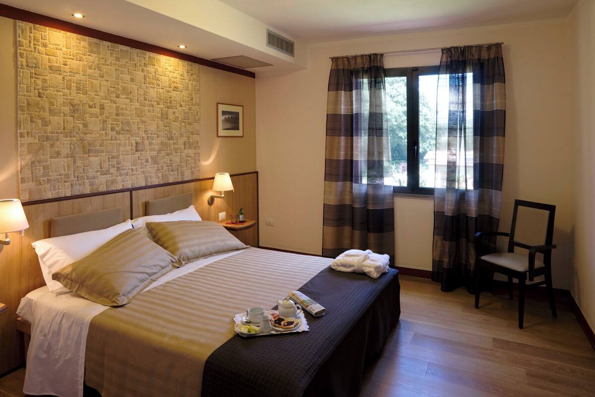 Habitación de hotel con pared de piedra, cama con cabecero, mesita de noche con lámpara, una silla y ventana con cortinas.