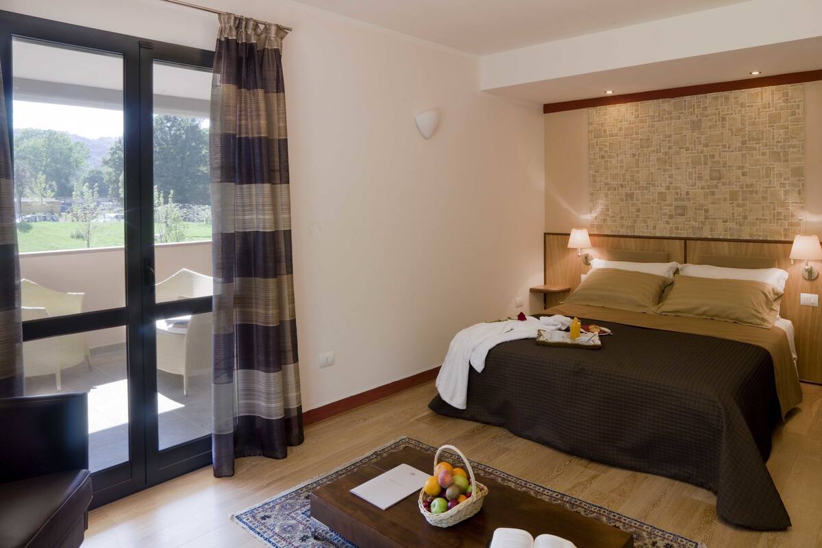 Habitación de hotel con cama, lámparas de noche, mesita de noche, ventana con cortinas y vistas a zonas verdes, además de una pequeña mesa con frutas.