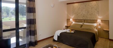 A hotel room with a bed, bedside lamps, a nightstand, a window with curtains, a view of greenery, and a small table with fruits.